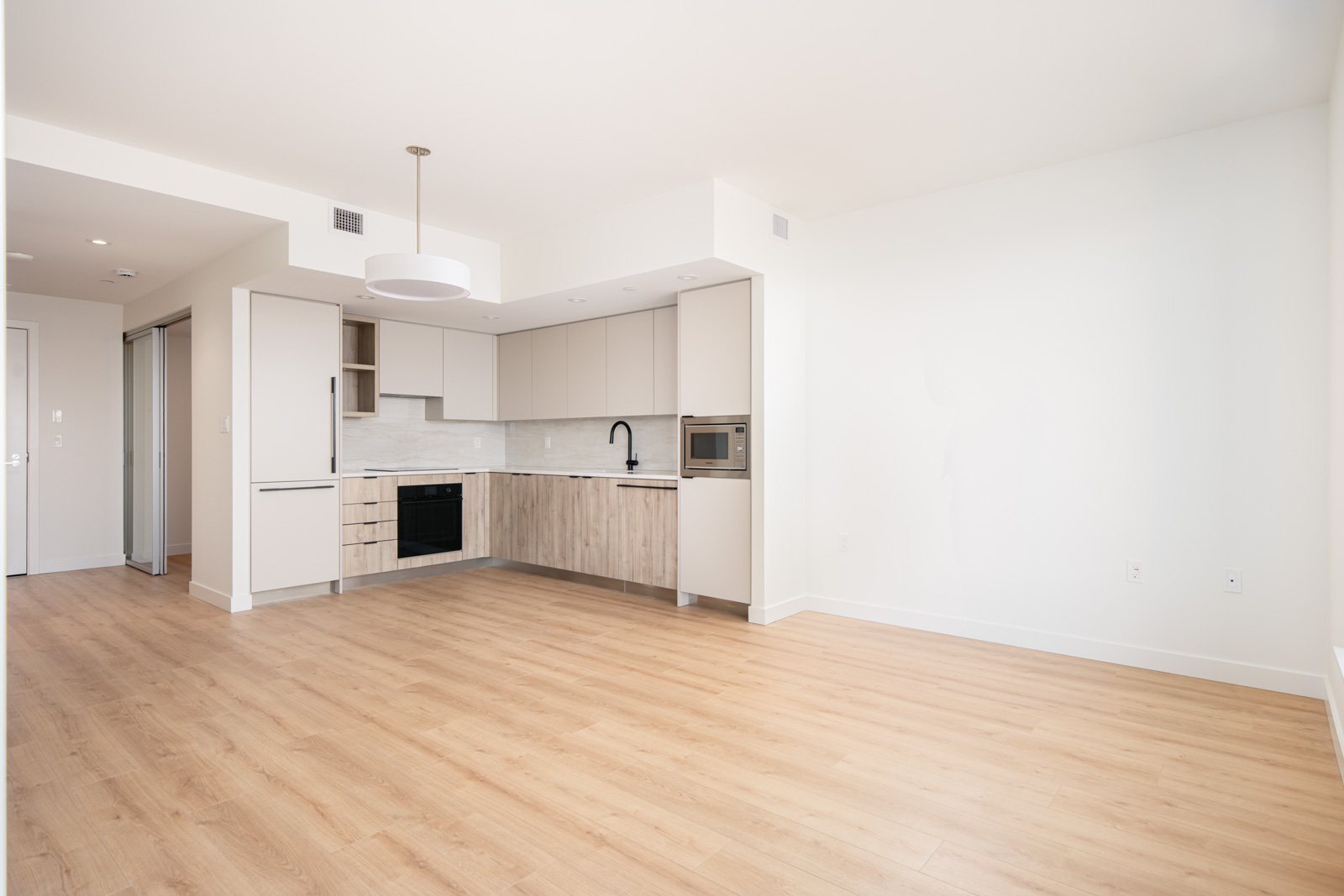 Modern, unfurnished apartment with light wood floors, white walls, and a minimalist kitchen featuring built-in appliances and light cabinetry.