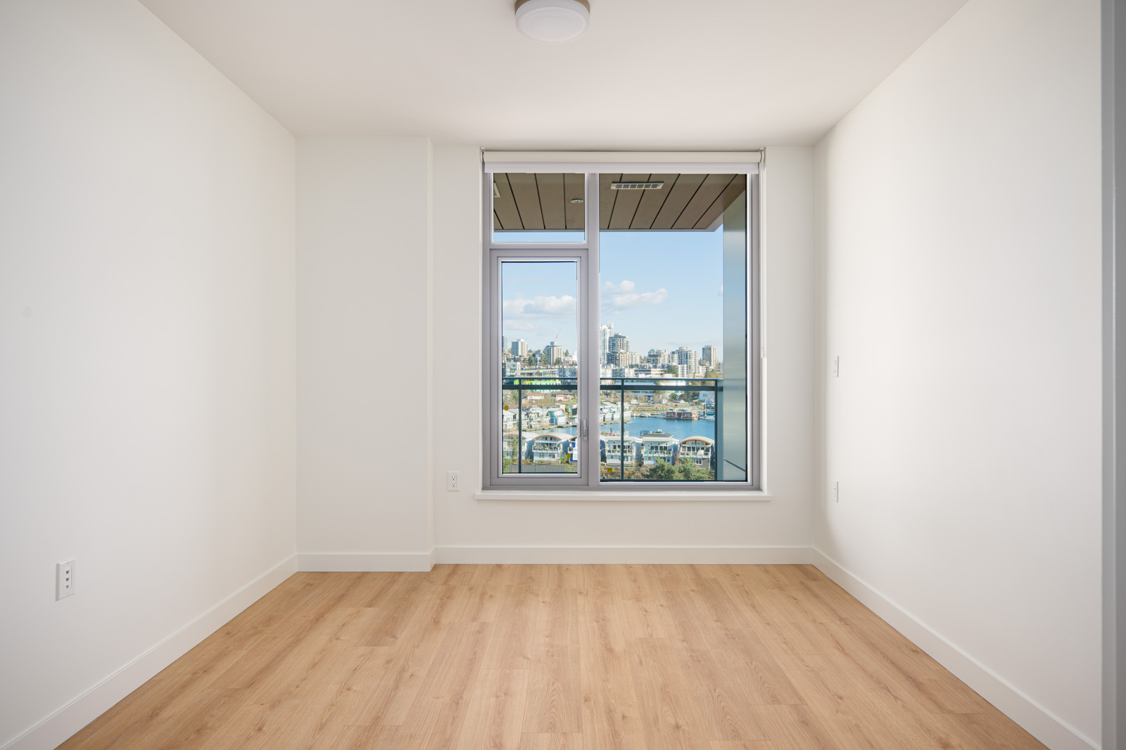 Empty, unfurnished room with light wood flooring, white walls, and a large window offering a city skyline and waterfront view.