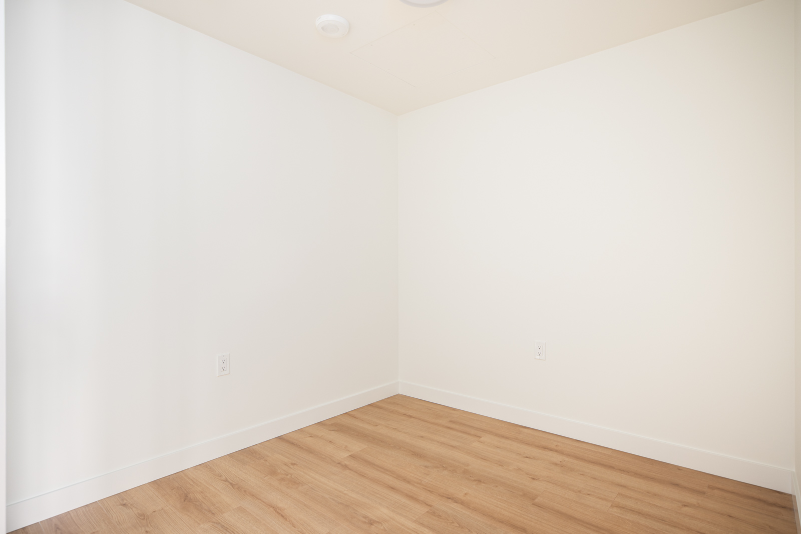 Empty room with white walls, light wood flooring, electrical outlets, and ceiling fixtures, shown from a corner perspective.