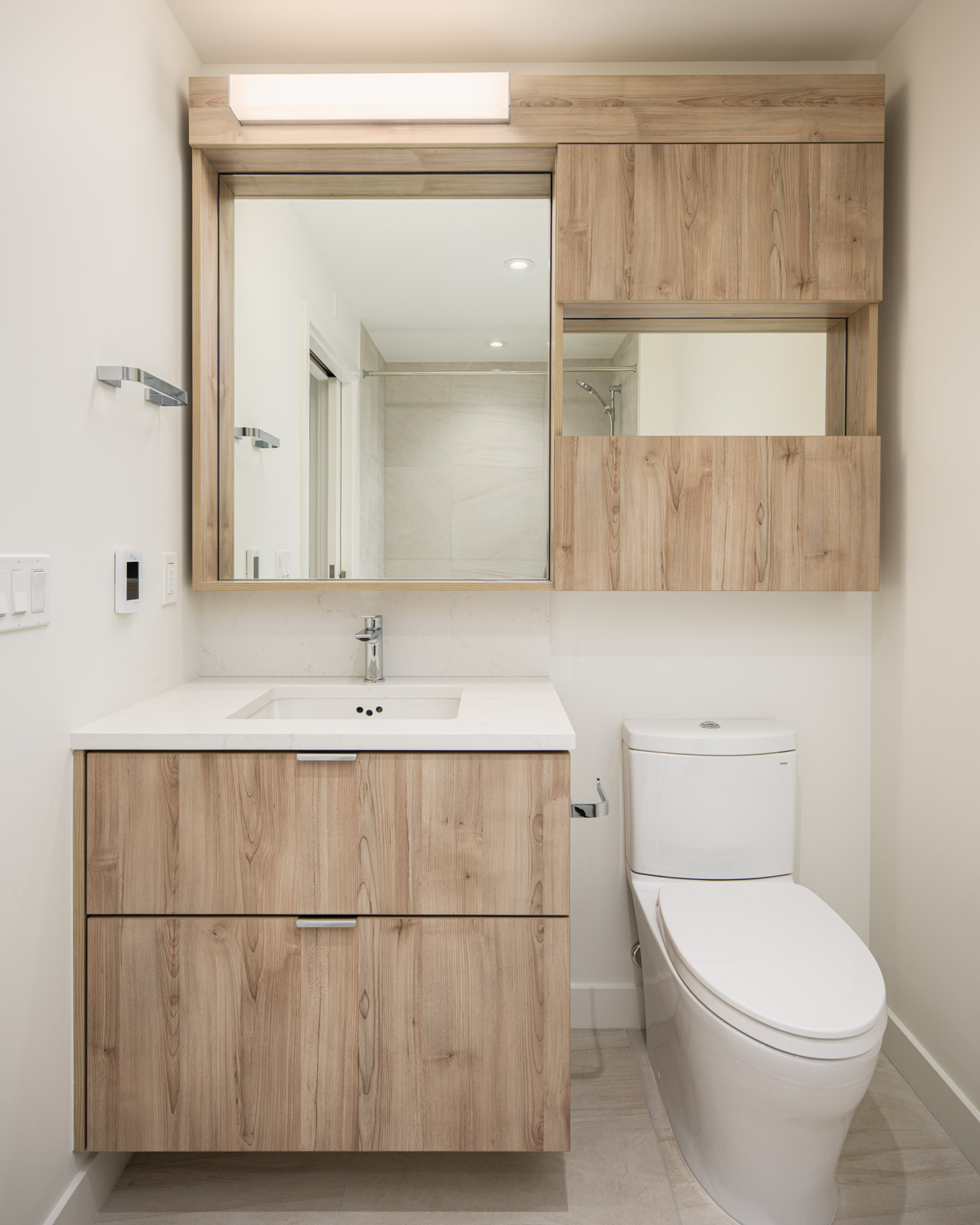 Modern bathroom with light wood vanity and cabinets, white countertop and sink, wall-mounted mirror, and white toilet on beige tiled floor.