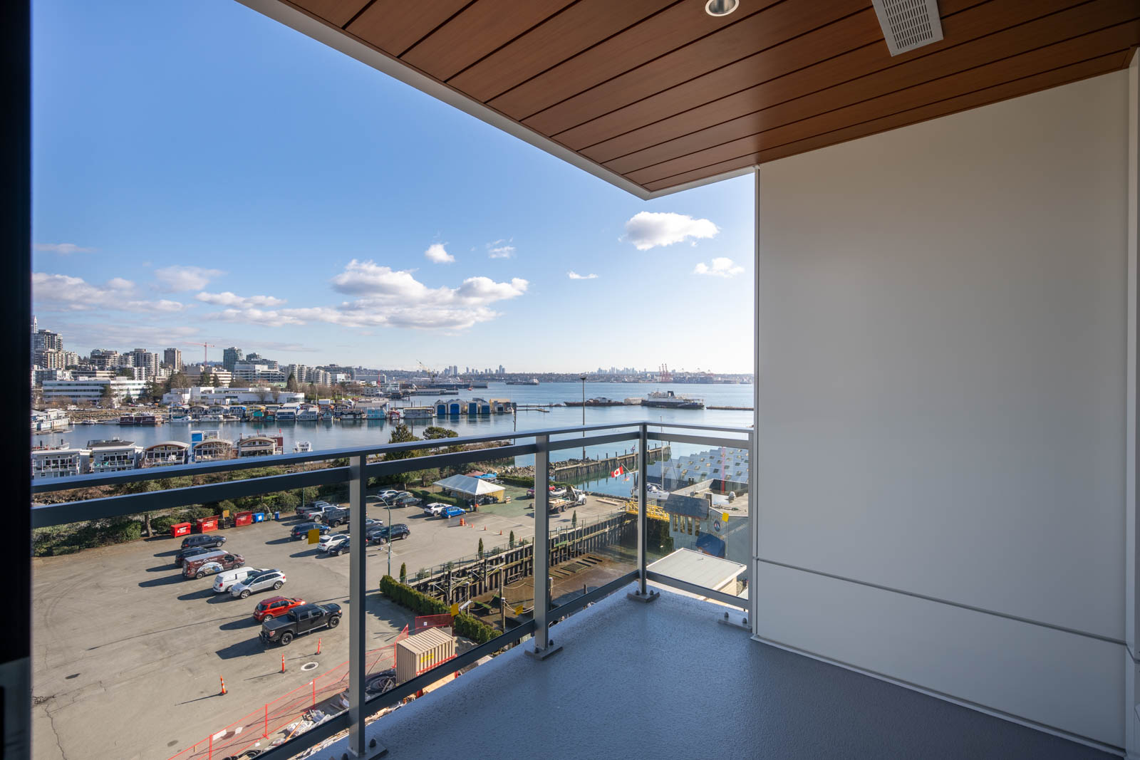 View from a balcony overlooking a parking lot, industrial buildings, and a harbor with boats, with a city skyline and blue sky in the background.