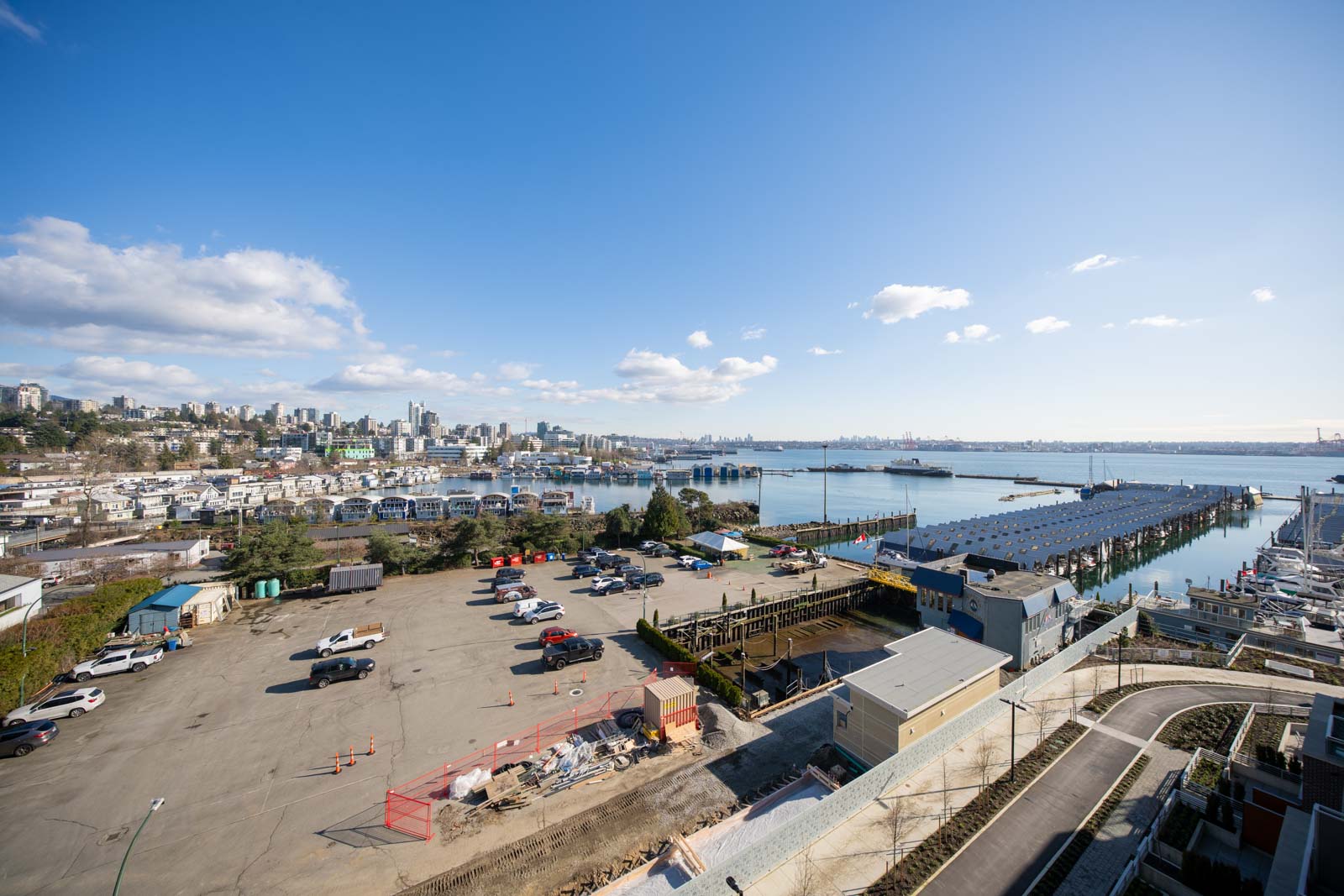 A waterfront industrial area with parked vehicles, warehouses, a pier, and a city skyline in the background under a clear blue sky.