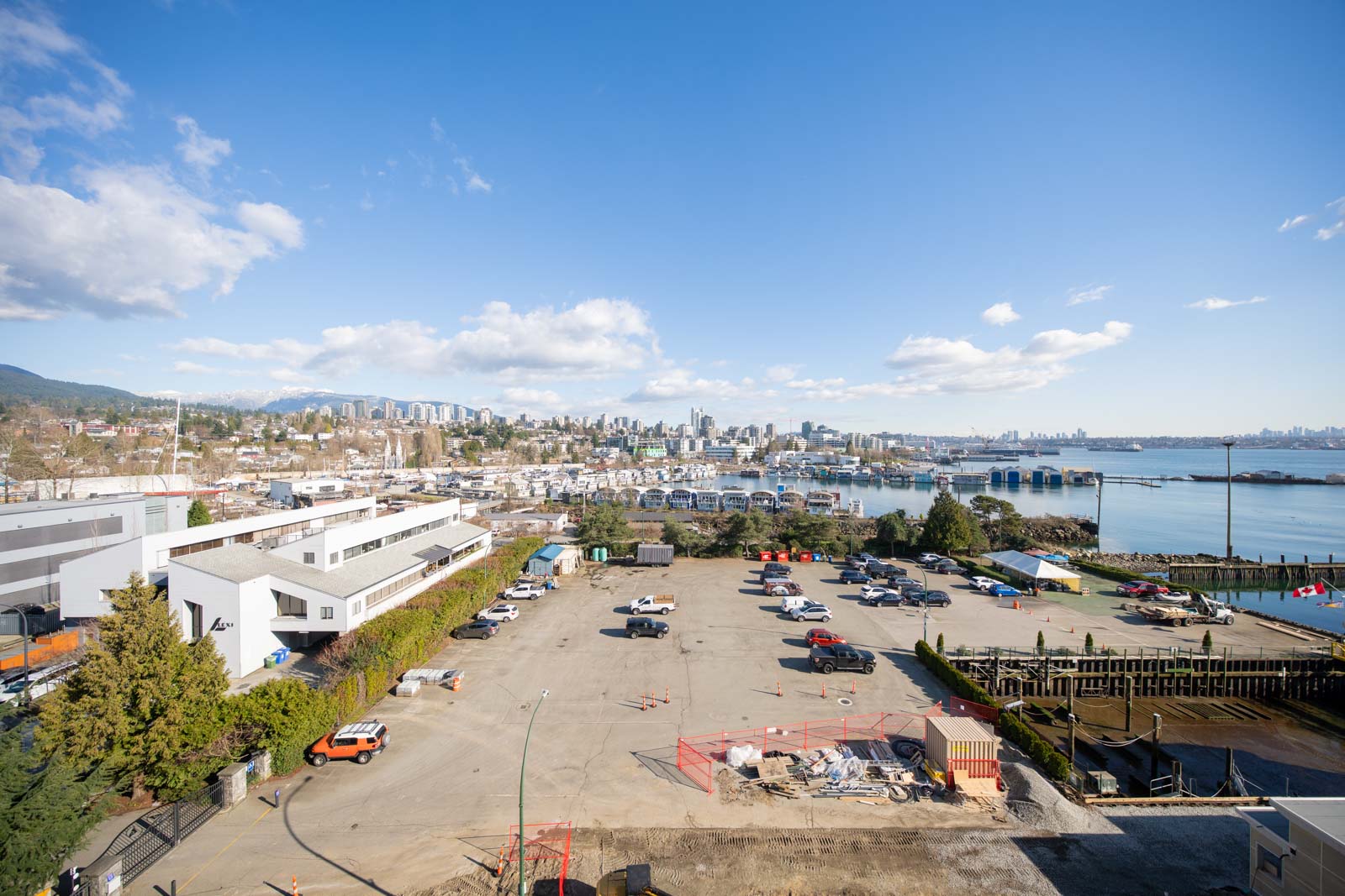 A wide view of a waterfront industrial area with parked vehicles, buildings, and a city skyline in the background under a clear blue sky.