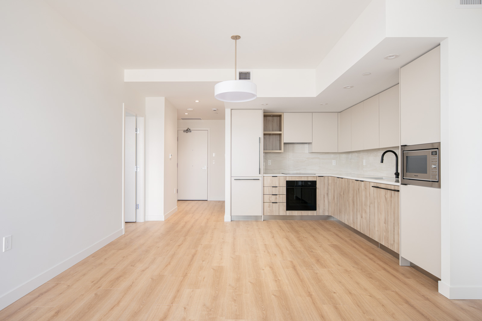 Modern empty apartment with light wood flooring, white walls, and a kitchen featuring built-in appliances, light cabinetry, and a ceiling light fixture.