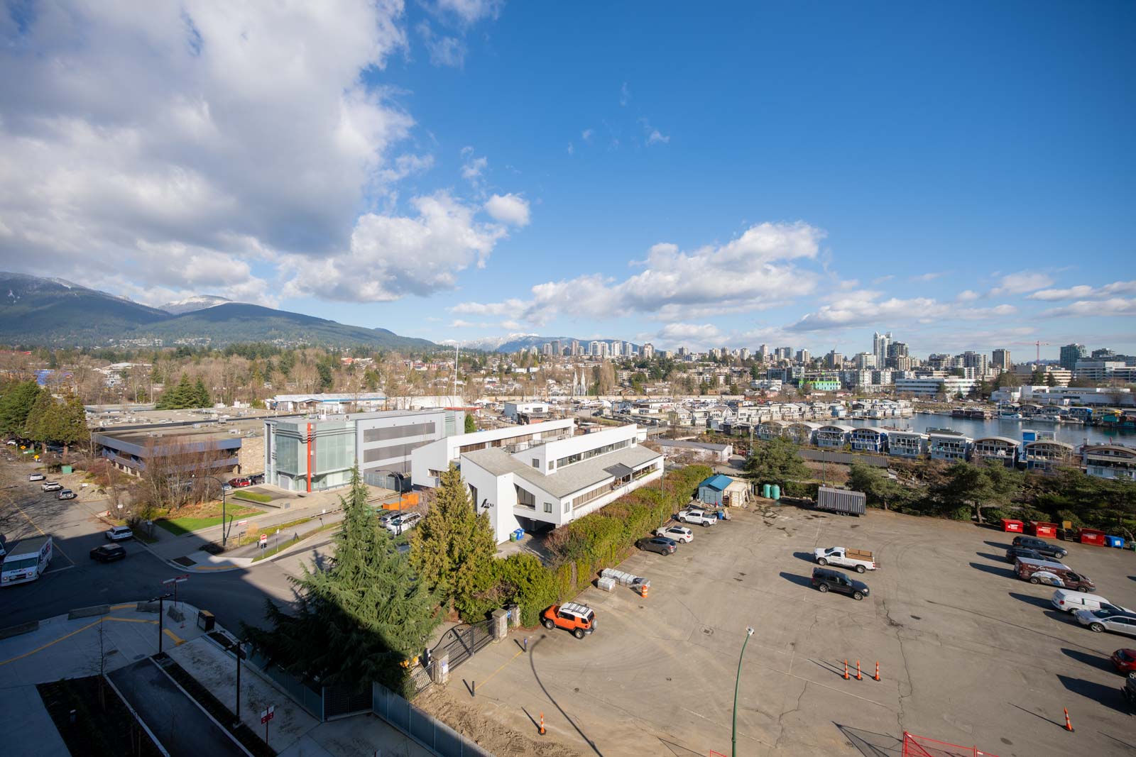 Aerial view of a city with buildings, parking lots, vehicles, and a marina, set against a backdrop of mountains and a partly cloudy blue sky.