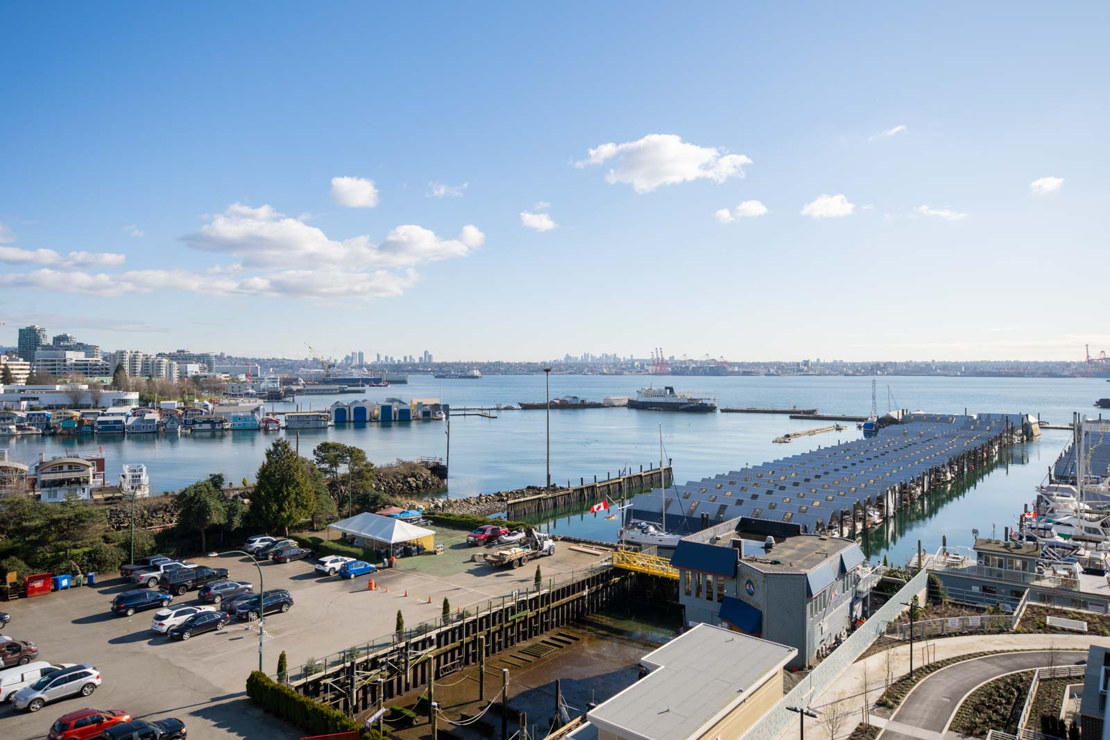 A waterfront marina with parked cars, several boats, dock buildings, and a city skyline in the distance under a clear blue sky.