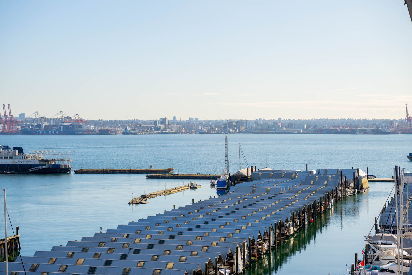 A marina with covered boat slips in the foreground, a ferry on the left, and an industrial port and cityscape across the water under a clear sky.