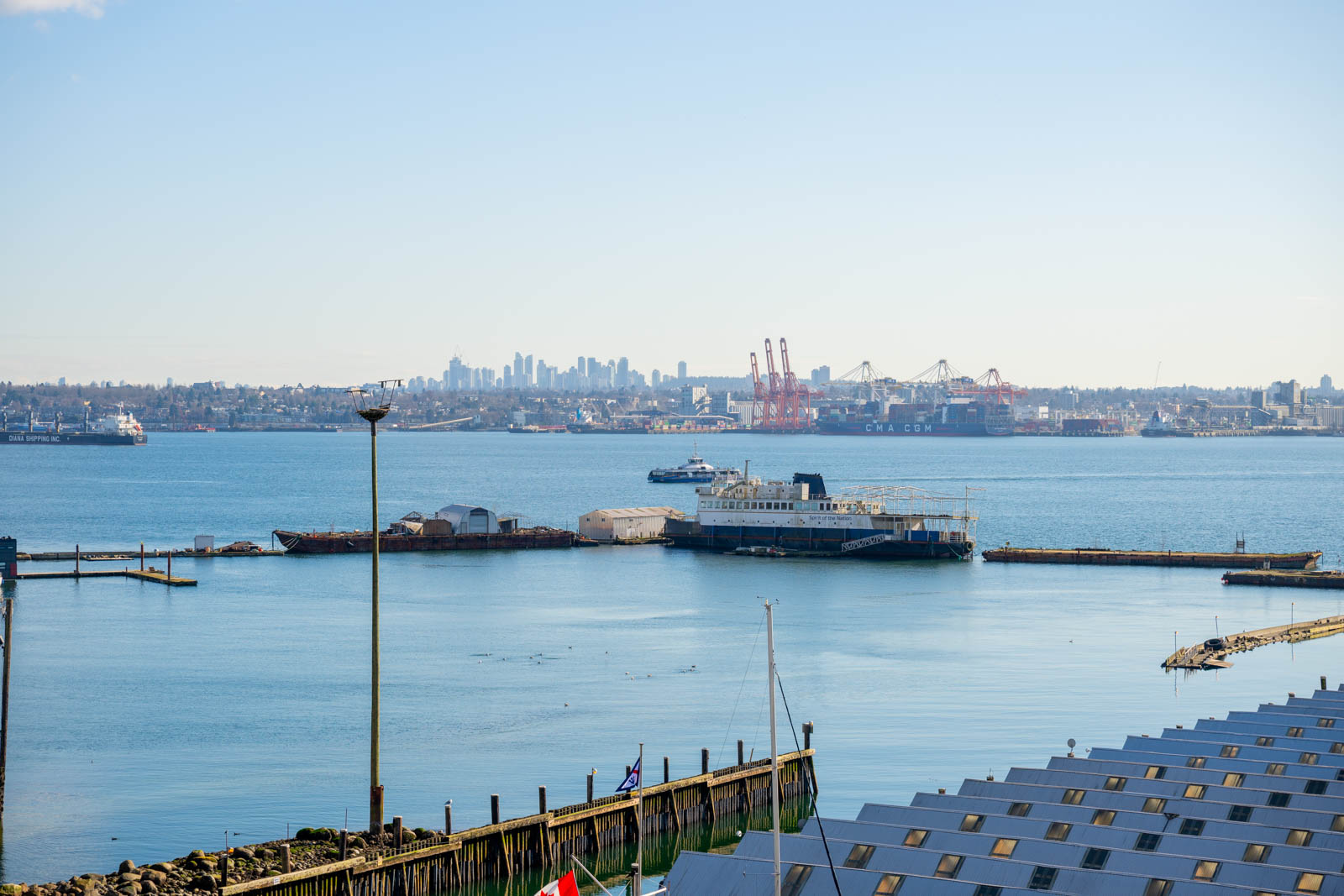 A waterfront scene with boats docked at a marina, shipping cranes in the background, and a distant city skyline under a clear sky.