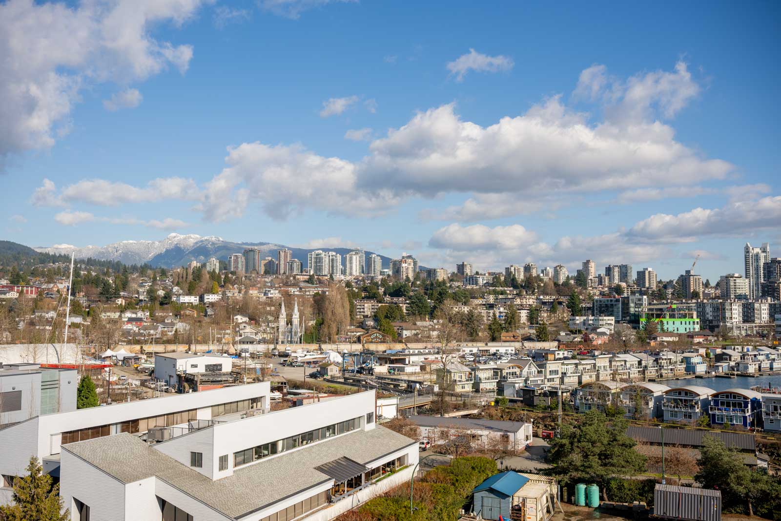 A cityscape with residential buildings in the foreground, high-rise buildings in the distance, and mountains under a partly cloudy blue sky.