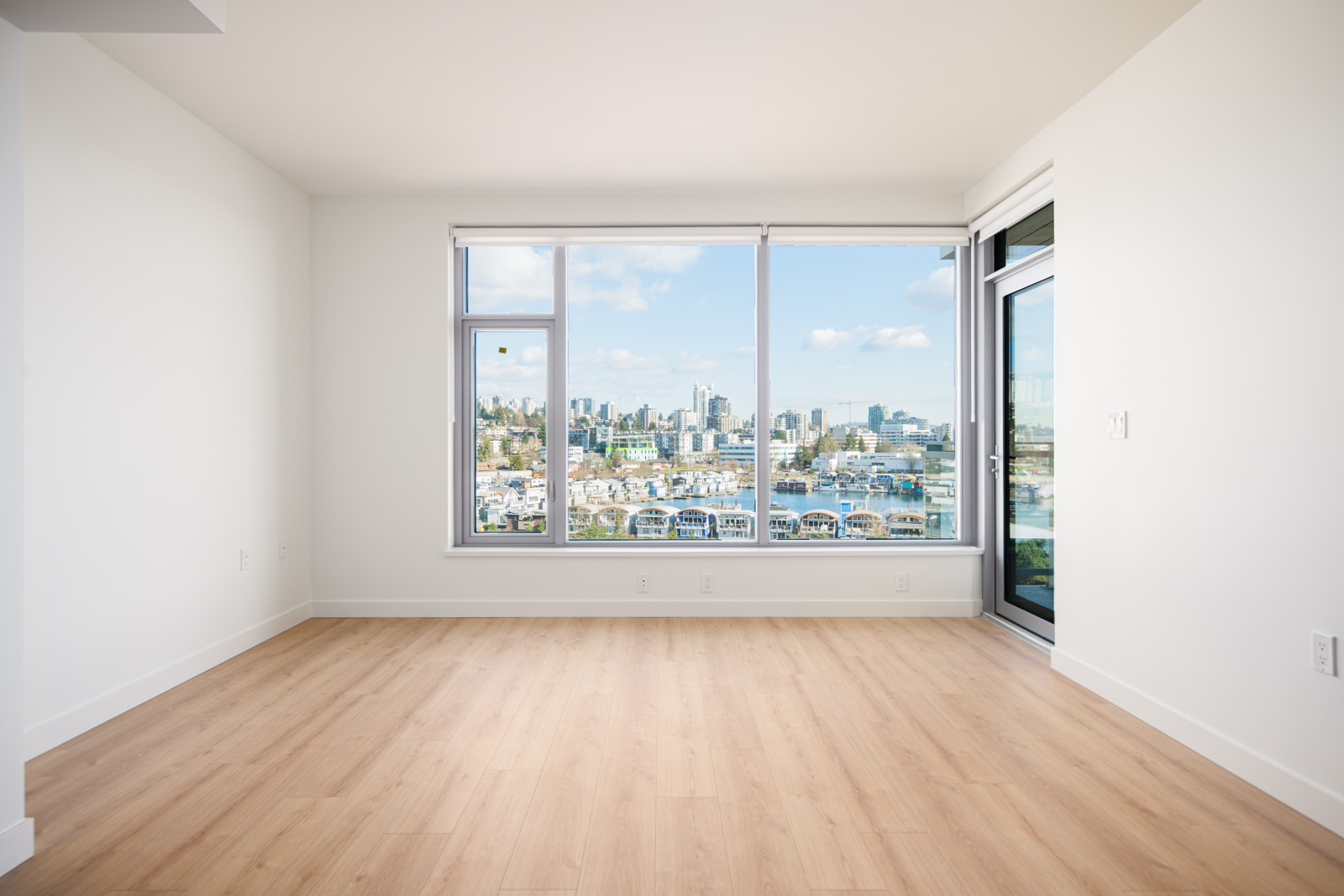Bright, empty room with light wood flooring, large window, and balcony door overlooking a city skyline and marina under a partly cloudy sky.