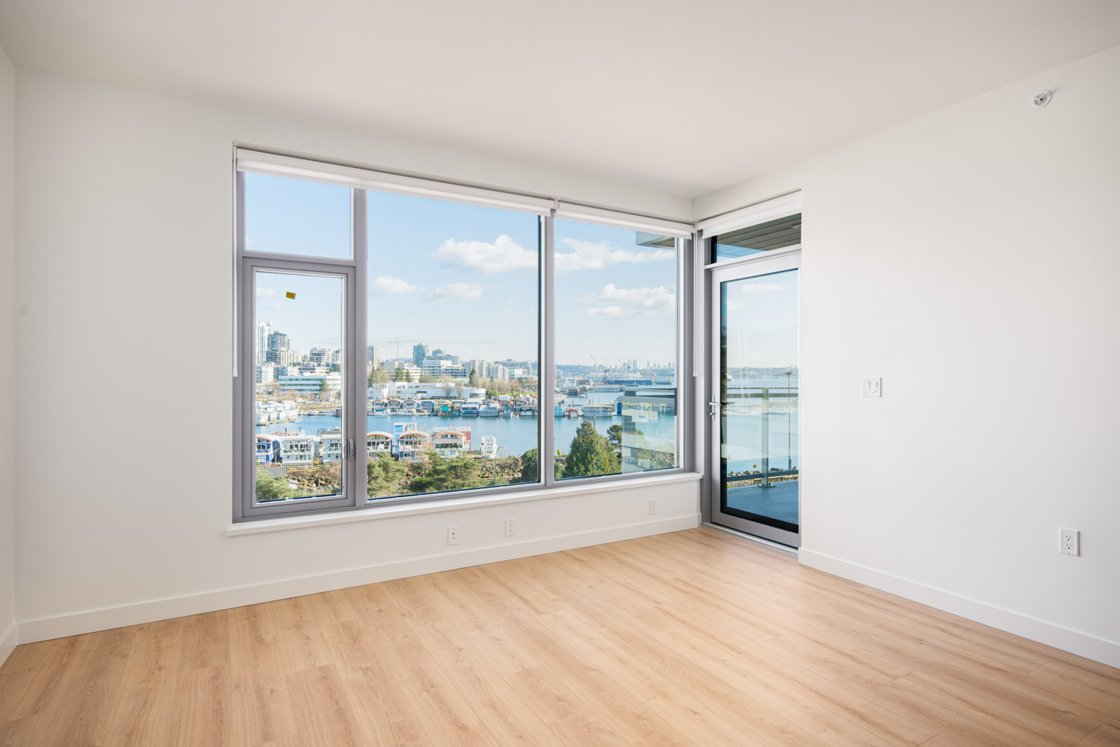 Unfurnished room with large windows and glass door, light wood flooring, and a city waterfront view under a blue sky.