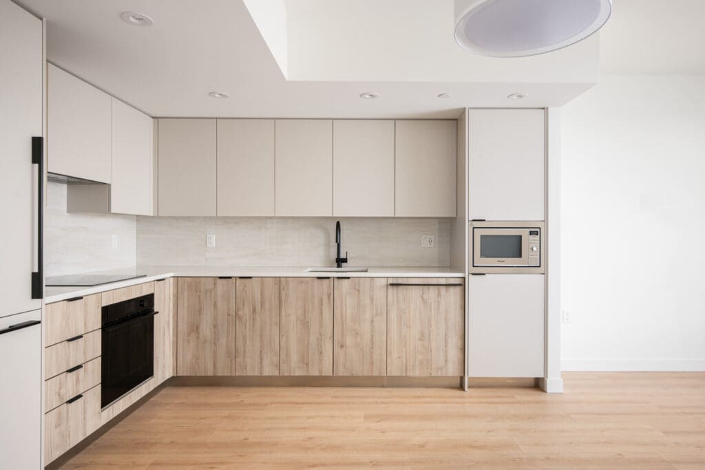 Modern kitchen with light wood lower cabinets, white upper cabinets, built-in oven, microwave, black sink fixtures, and light wood flooring. The kitchen is clean and minimalistic.