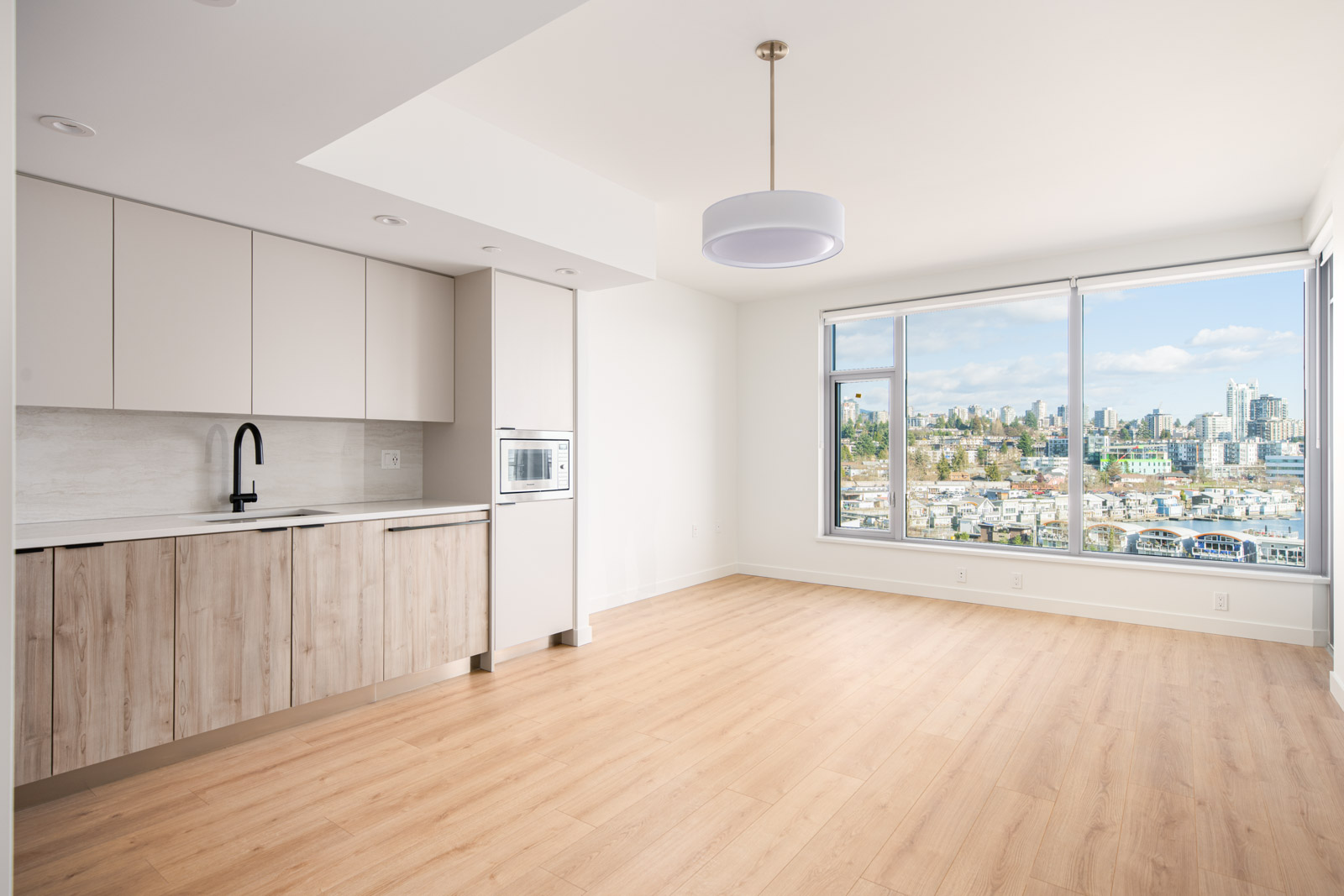 Modern, unfurnished apartment with light wood floors, minimalist kitchen, large windows, cityscape and marina views, and a ceiling light fixture.