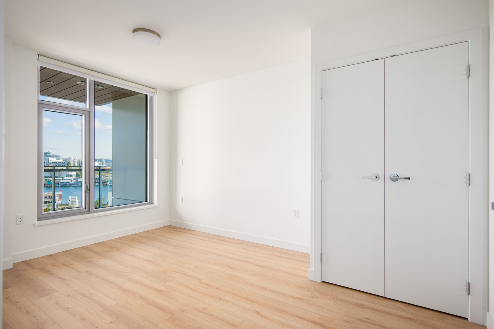 Empty room with light wood flooring, white walls, a double-door closet, and a large window overlooking a cityscape and waterfront.