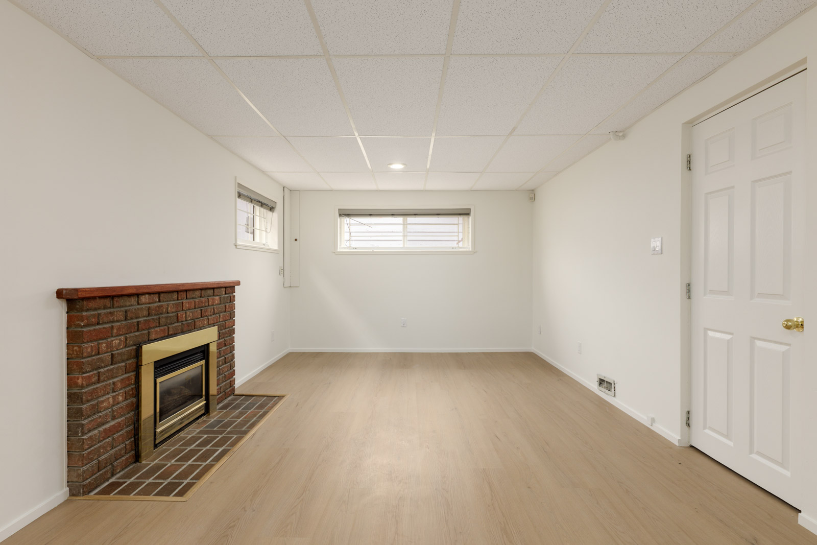 Unfurnished basement room with light-colored walls, wood-style flooring, a brick fireplace, and a small window letting in natural light.