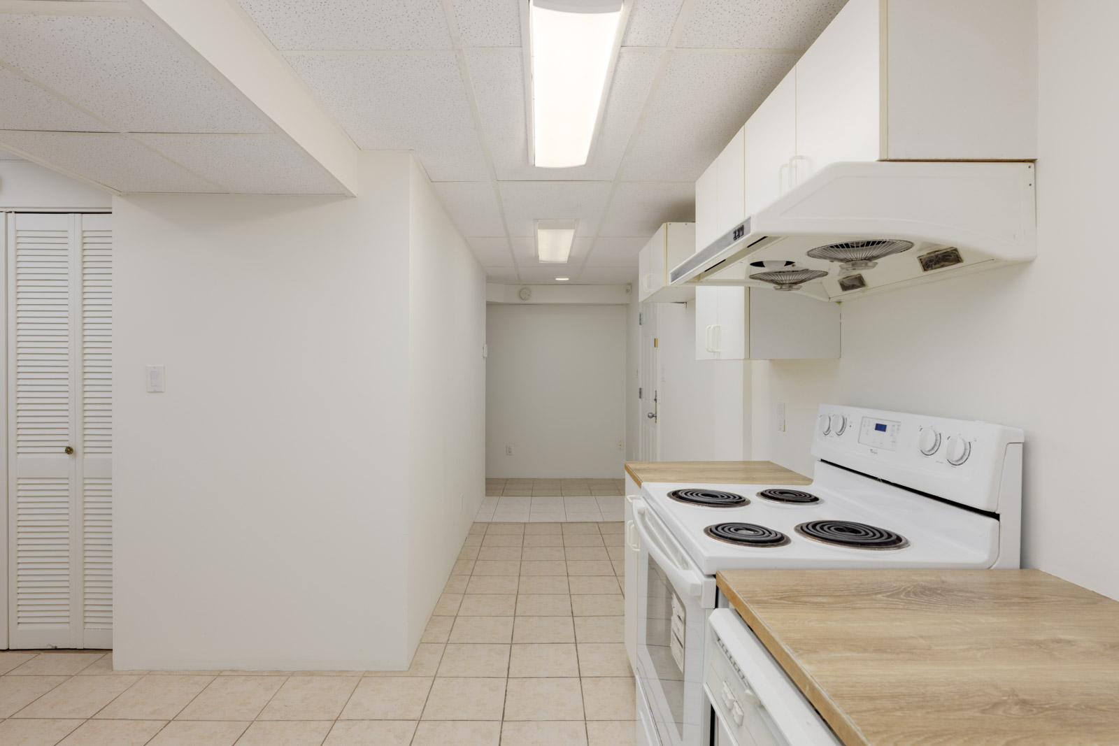 A narrow kitchen with white walls, electric stove, range hood, wooden countertops, and tile flooring, leading to a small hallway with closed closet doors and ceiling lights.