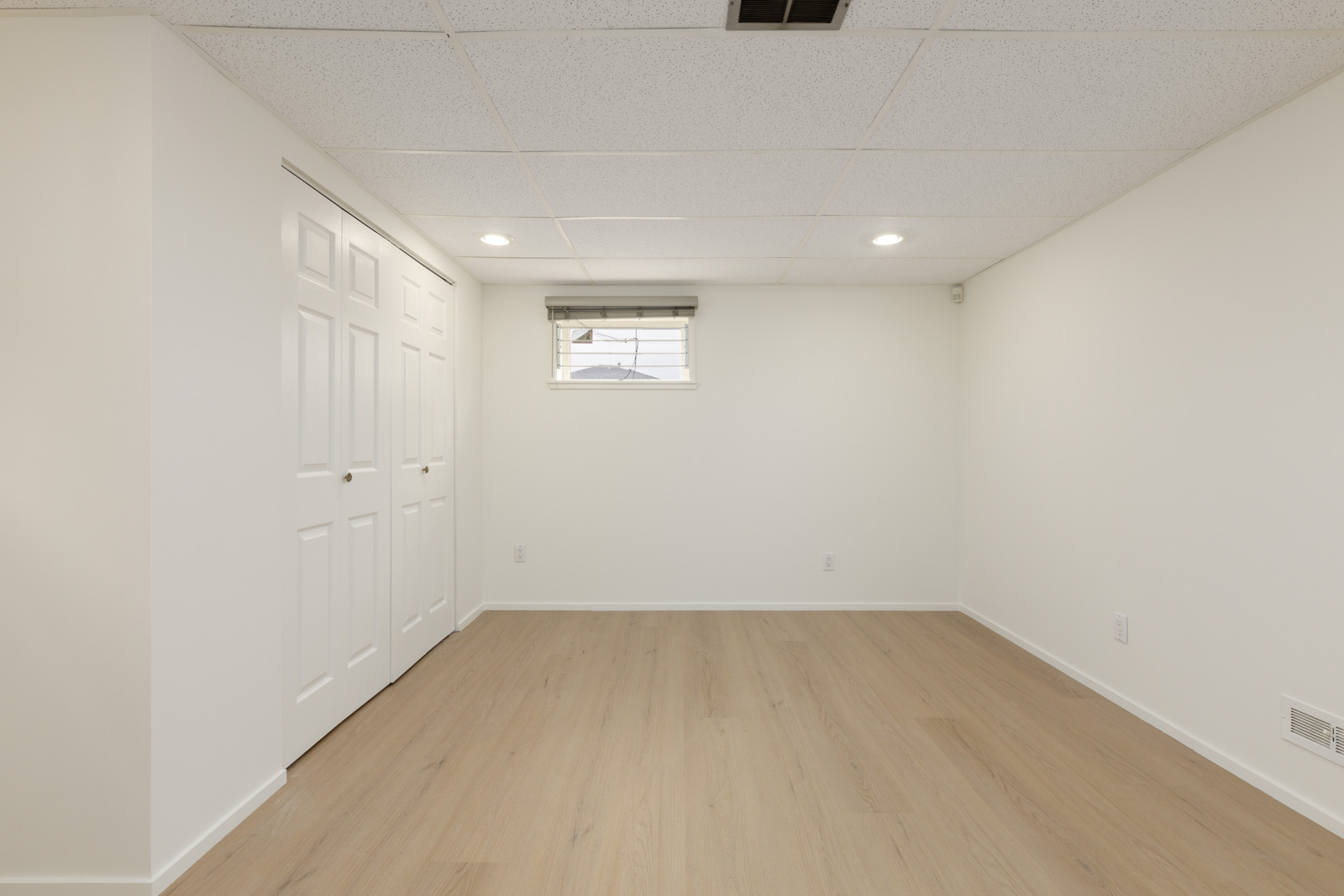 Empty room with light wood flooring, white walls, a small window with blinds, recessed ceiling lights, and double closet doors.