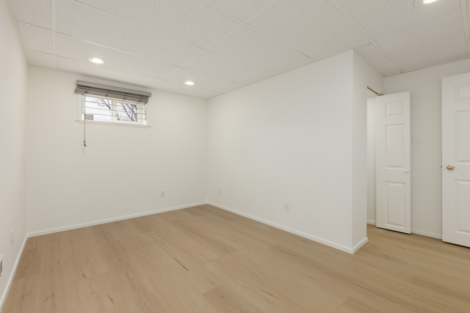 Empty room with light wood flooring, white walls, a small window with blinds, recessed ceiling lights, and two white doors, one partially open.