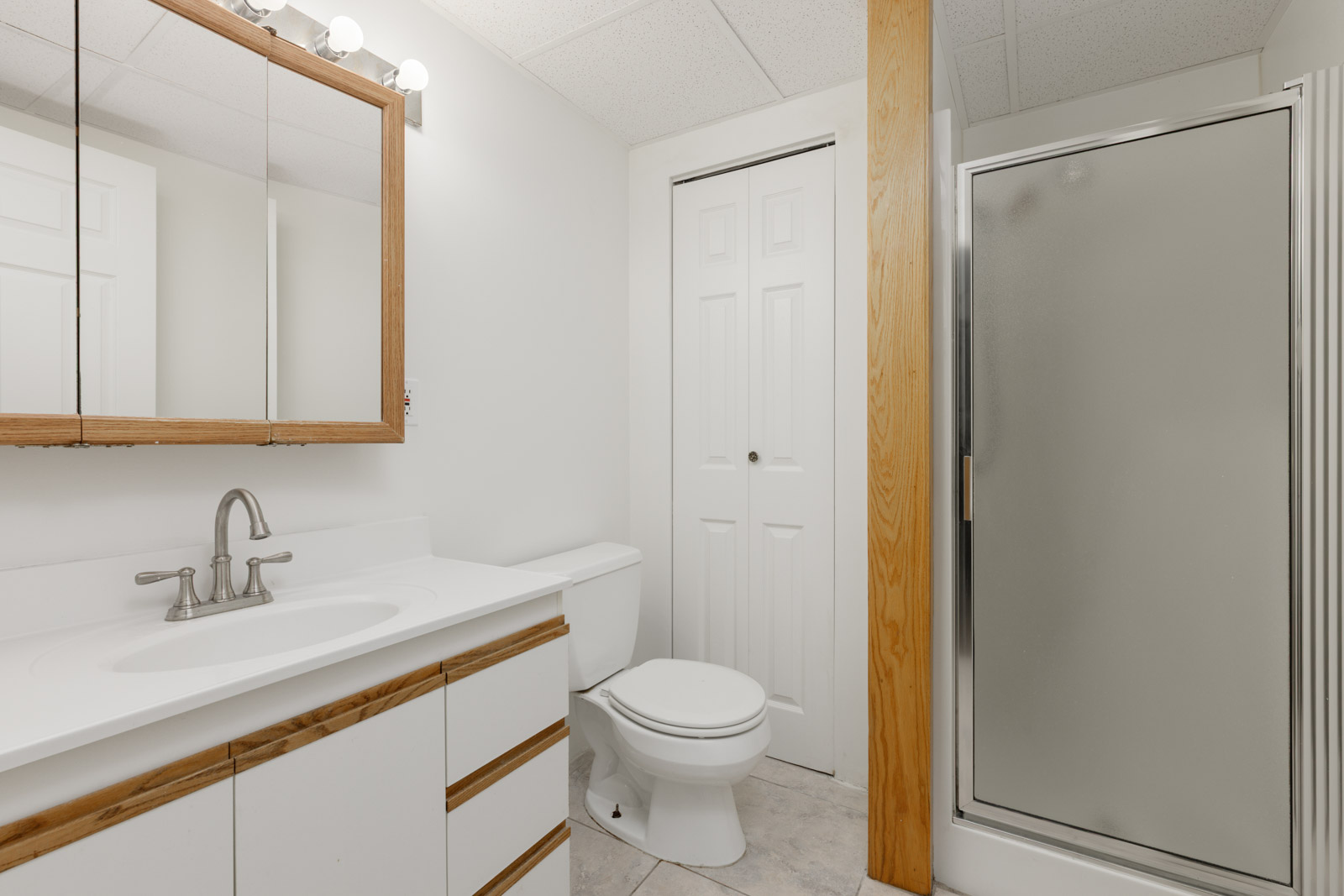 A bathroom with a sink, mirror, toilet, white cabinets, a frosted glass shower, and a closet with double doors.