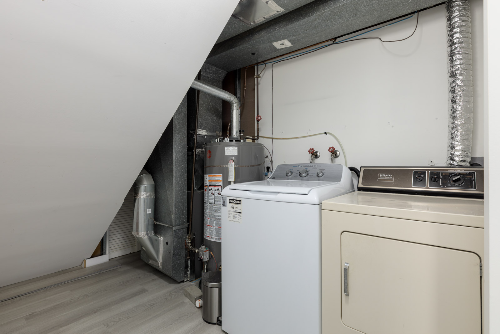 Laundry area with a white washing machine, beige dryer, water heater, vent pipes, and exposed ductwork against a slanted ceiling and white wall.