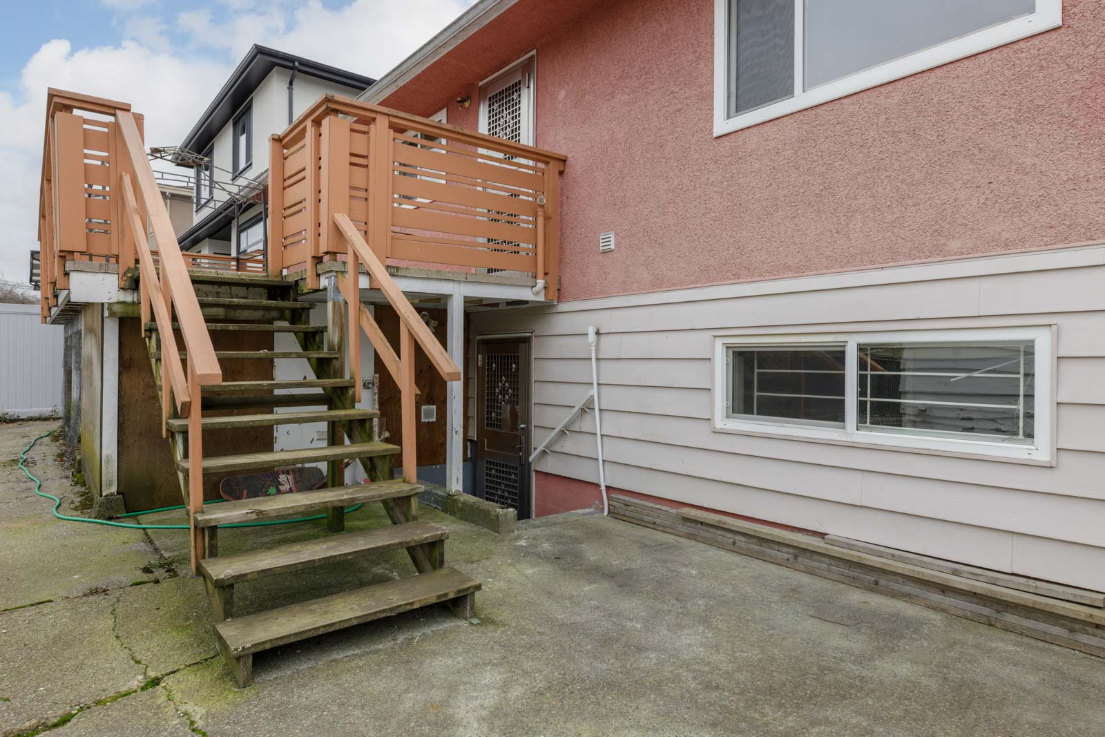 Outdoor wooden staircase leads to a second-floor entrance; ground-level door and window visible on stucco and siding exterior. Concrete patio in foreground.