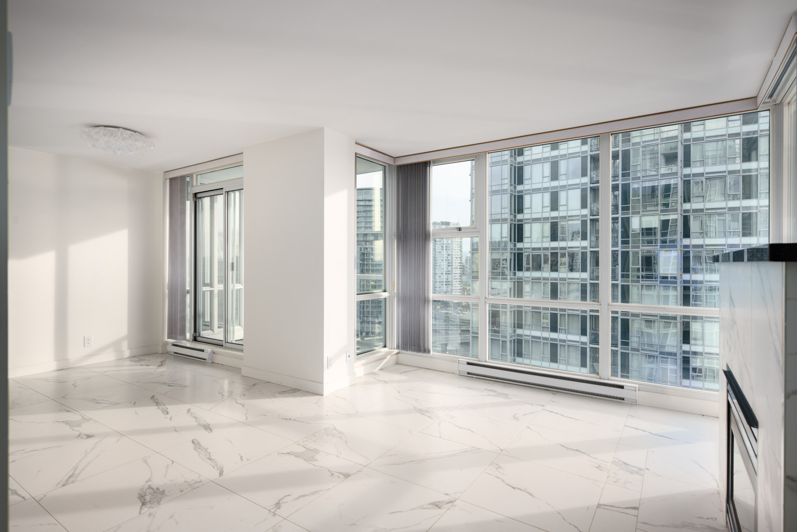 A bright, empty modern apartment room with large windows, marble tile flooring, and views of nearby high-rise buildings.