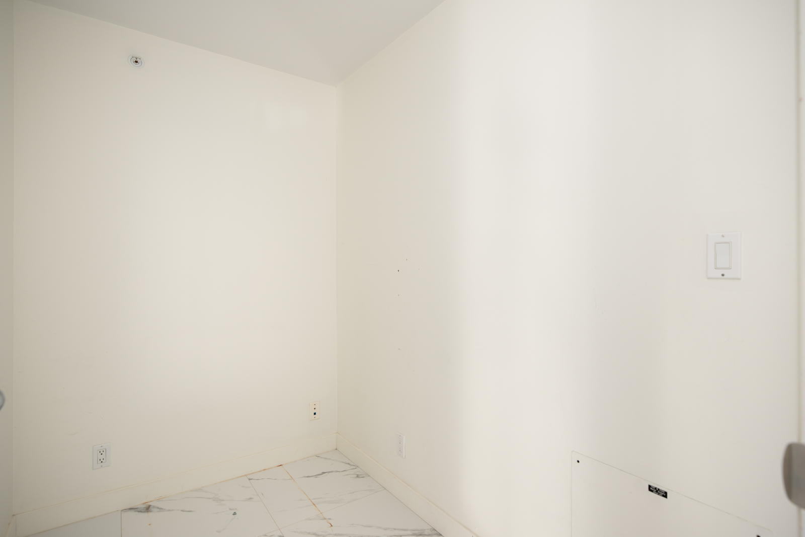Empty white room with tile flooring, a wall light switch, electrical outlets, and a small wall panel visible.