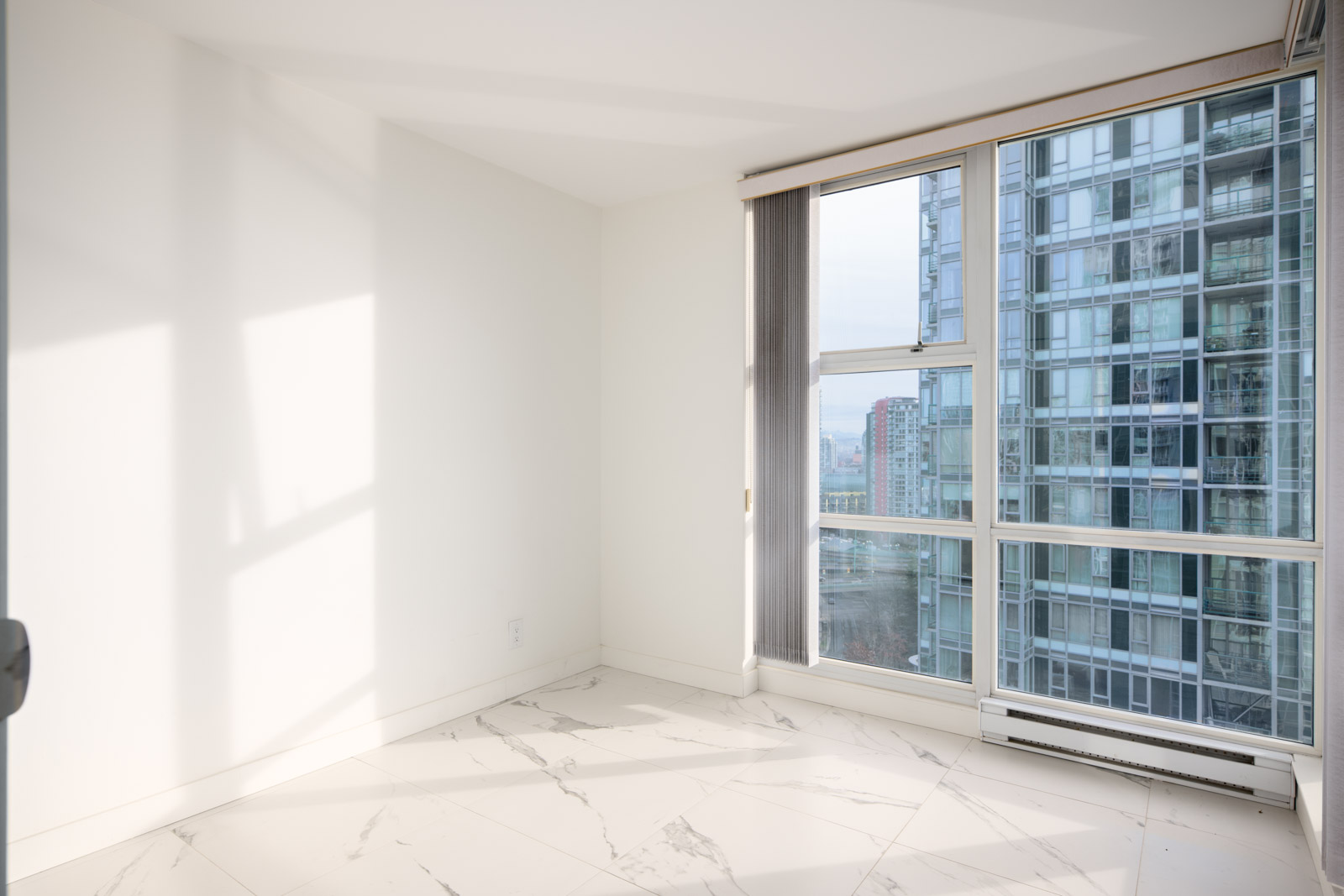 Empty, sunlit room with white walls, marble tile floor, and large windows overlooking modern high-rise buildings.