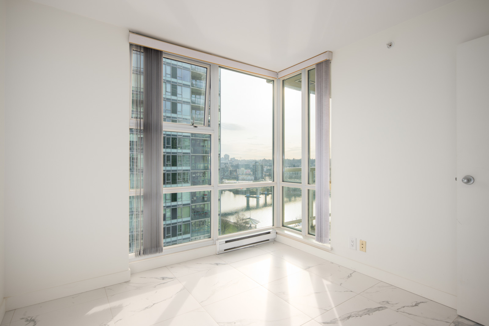 Bright, empty room with large corner windows, white walls, and marble tile flooring, overlooking city buildings and a river.