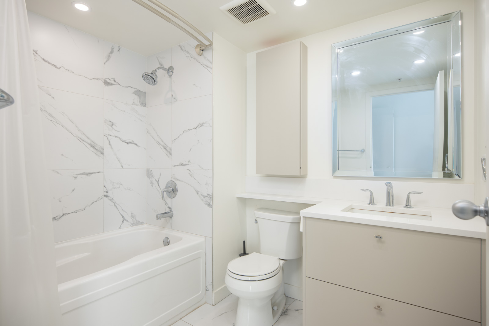 Modern bathroom with white marble tile in shower, beige vanity with sink, wall cabinet, toilet, large mirror, and overhead lighting.