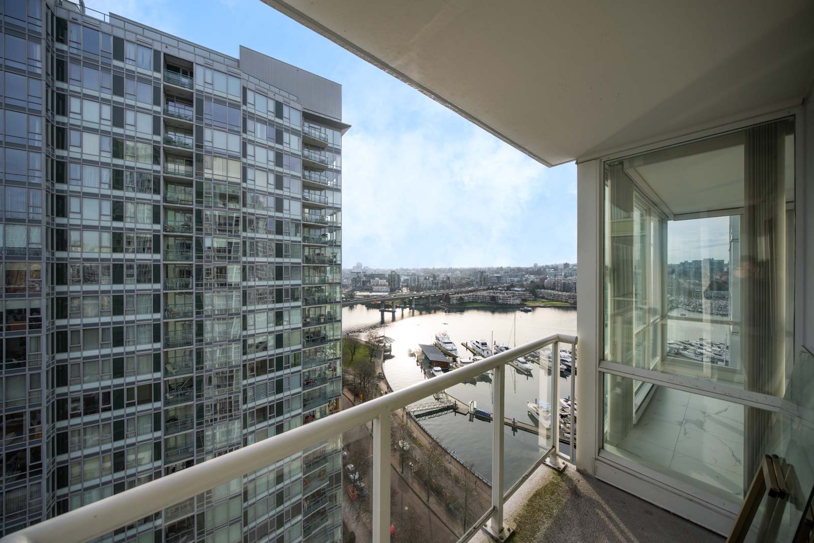 View from a high-rise balcony overlooking a river, marina with boats, and adjacent glass-clad residential building on a clear day.