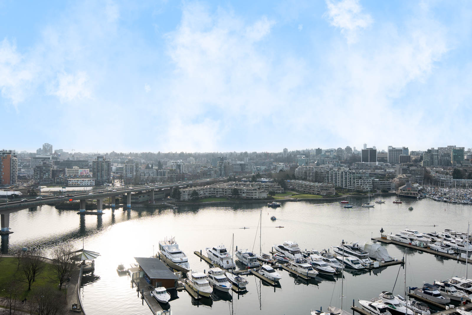 A marina with several docked boats on calm water, a bridge to the left, and a city skyline in the background under a partly cloudy sky.