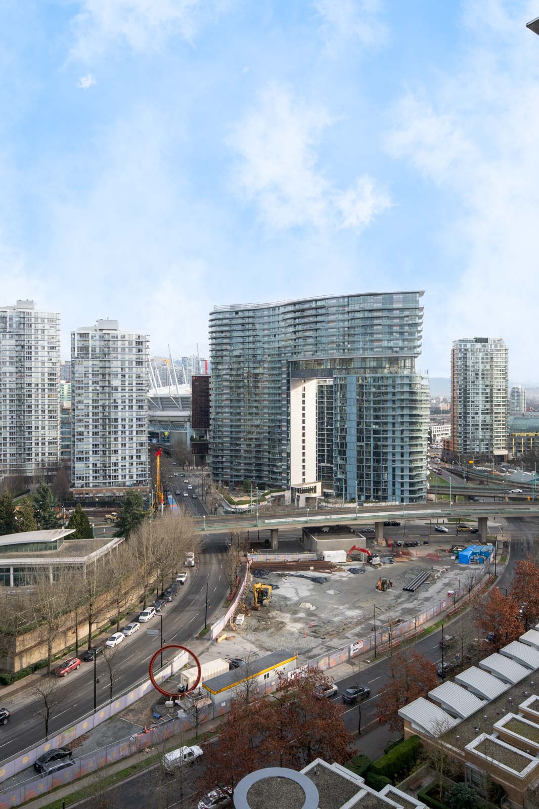 A cityscape featuring high-rise buildings, a construction site, and multiple roads with vehicles under a partly cloudy sky.