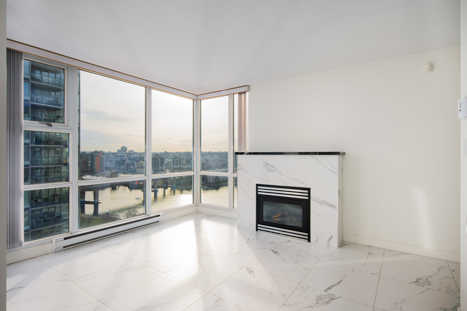 Modern, unfurnished corner apartment with large floor-to-ceiling windows, marble tile flooring, a gas fireplace, and a city and water view.
