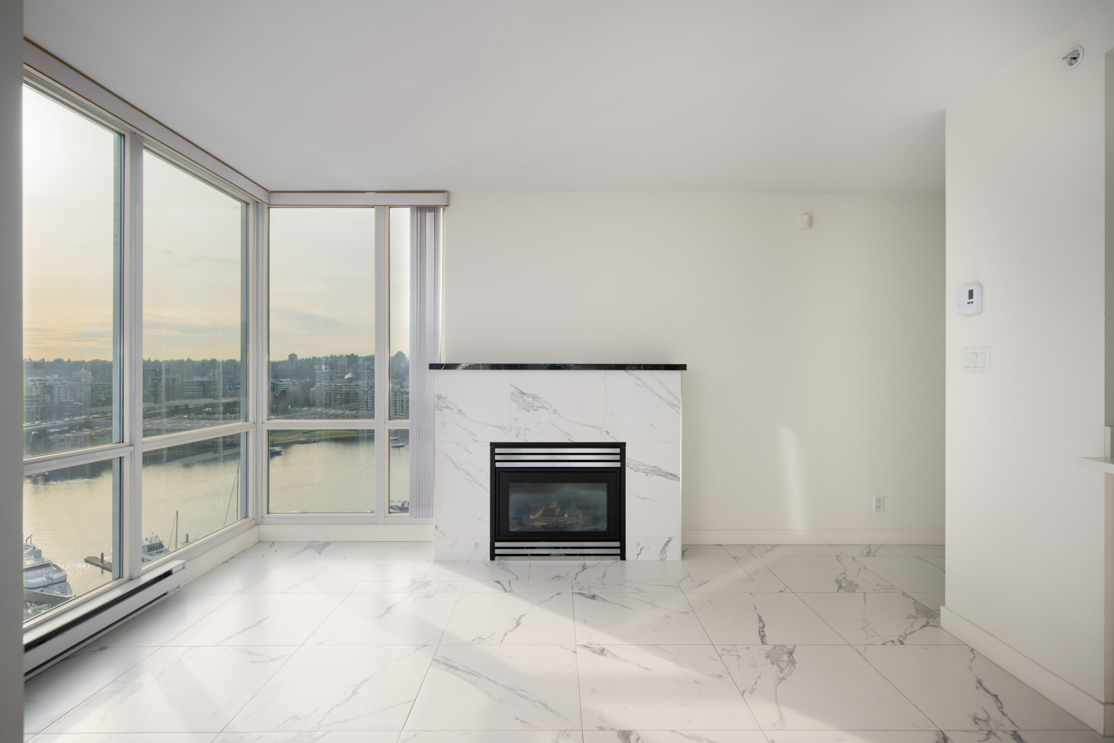 Bright, modern living room with large windows, marble tile flooring, and a marble fireplace overlooking a waterfront view.