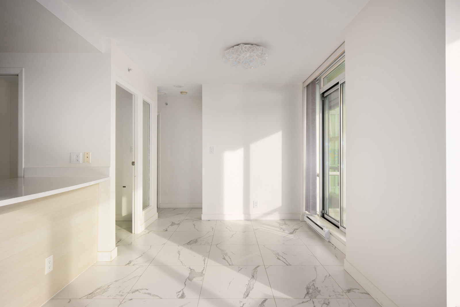 Bright, empty room with white walls, large tile flooring, sliding glass door, and modern light fixture. Sunlight streams through the window.