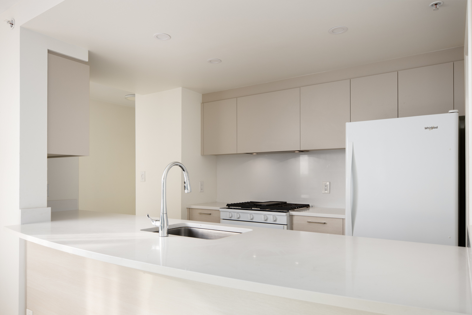 Modern kitchen with a white countertop, built-in gas stove, overhead cabinets, and a white refrigerator. A stainless steel sink with a faucet is in the foreground.
