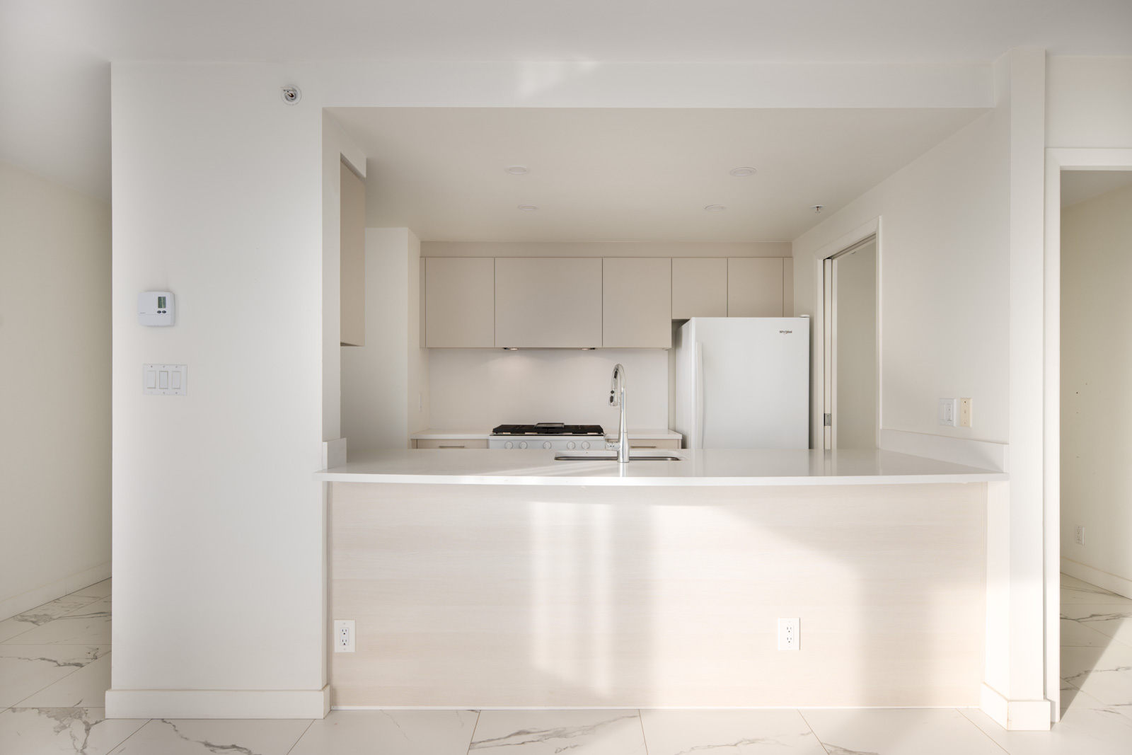 Minimalist kitchen with white cabinets, countertop, and appliances. Marble floor tiles and a central island with a sink are visible. The space is brightly lit.