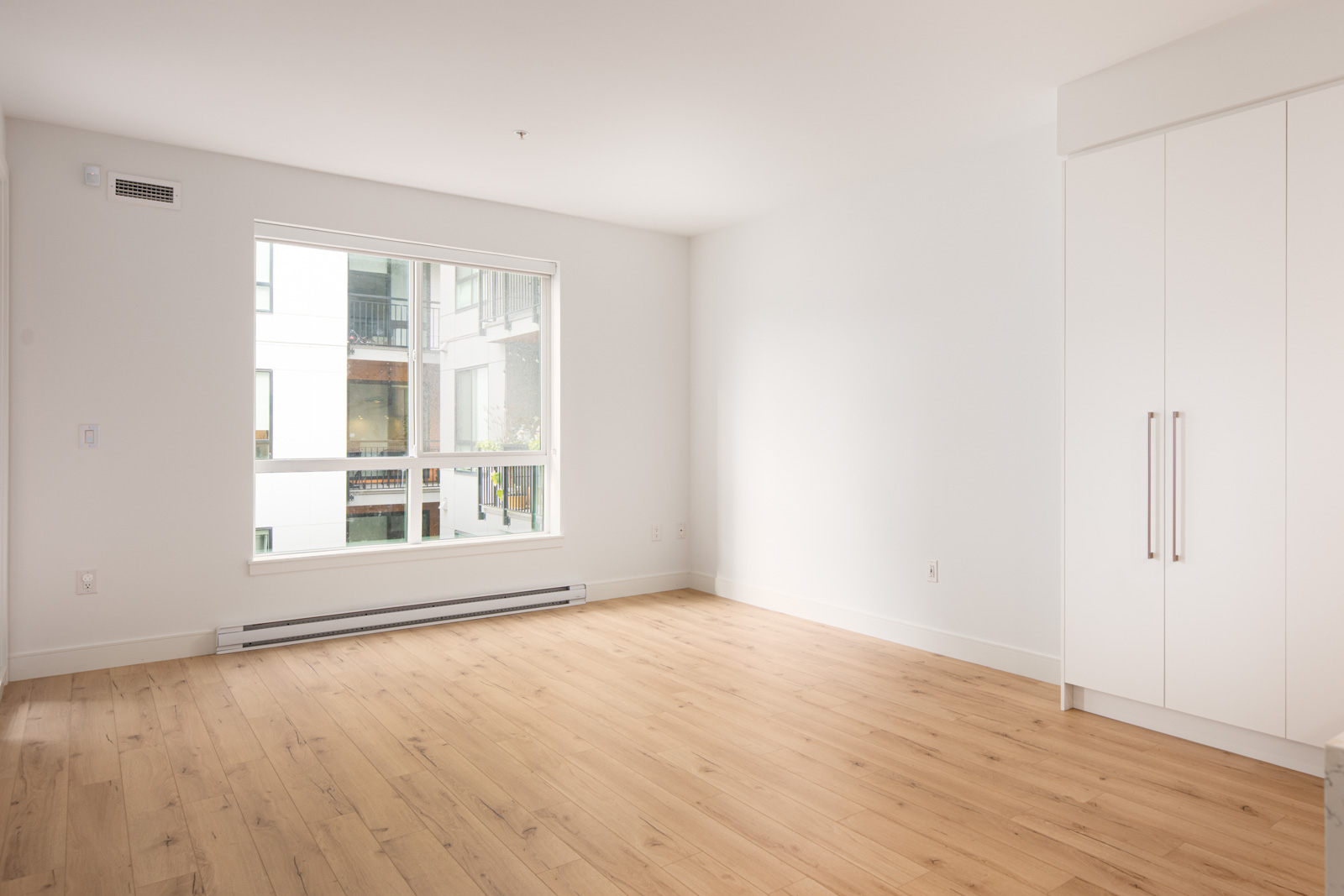 Empty, unfurnished room with light wood flooring, white walls, a large window, and built-in white closet. Exterior of nearby building visible through the window.