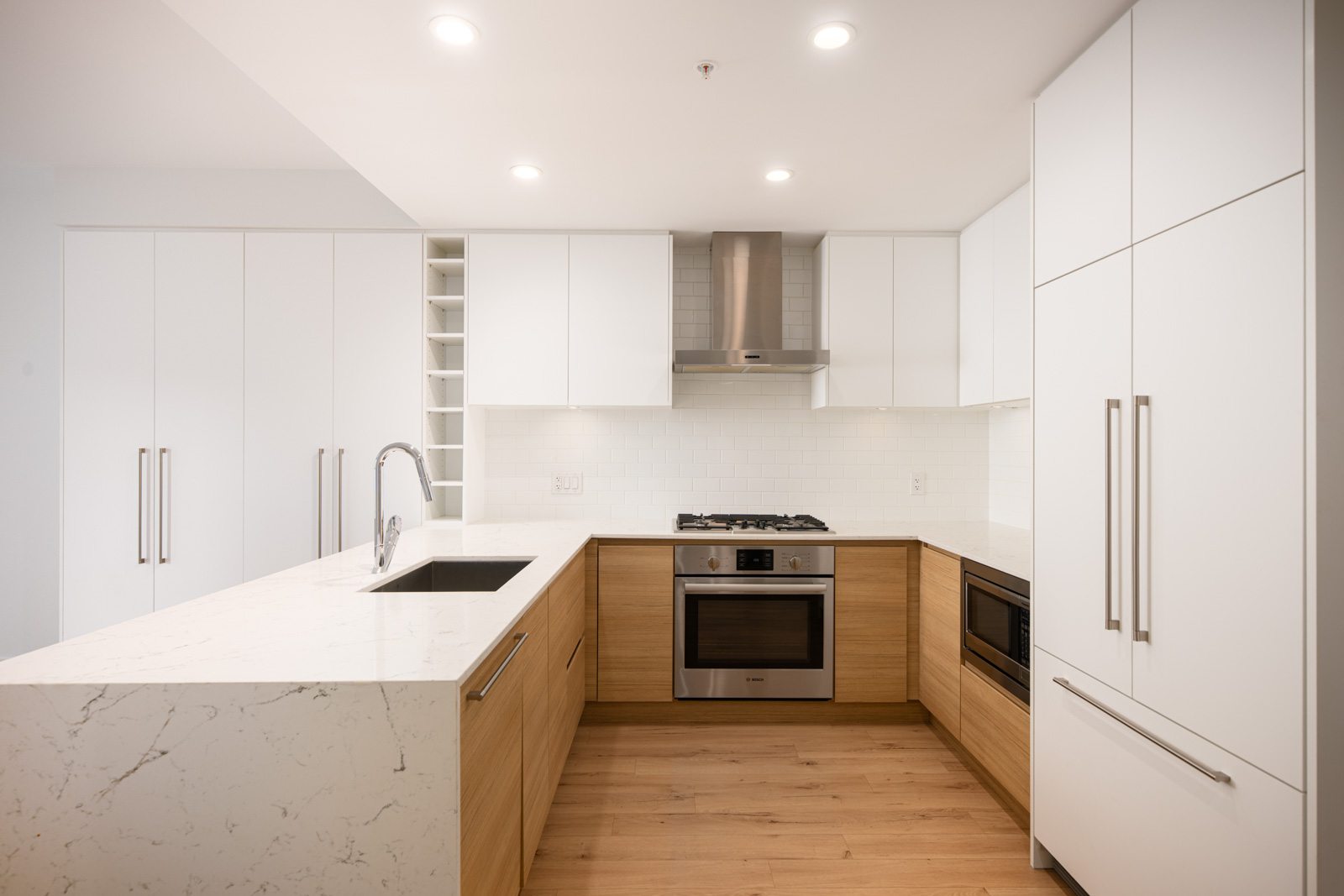 Modern kitchen with white cabinets, stainless steel appliances, built-in oven, gas cooktop, marble countertops, and light wood lower cabinets; bright lighting and minimalist design.