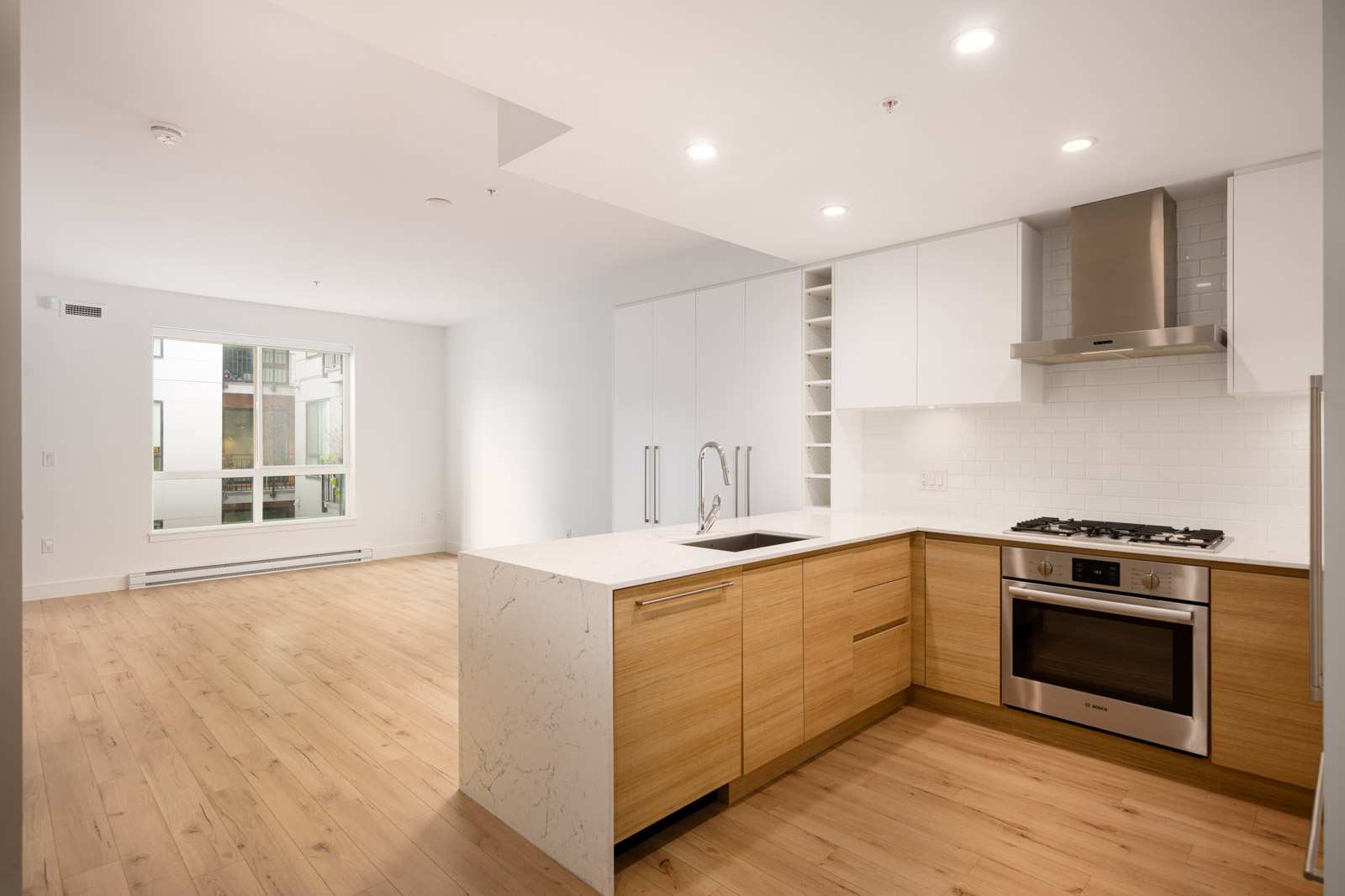 Modern open-plan kitchen with wood and white cabinetry, built-in oven, gas cooktop, and a marble island; adjacent to a bright living area with large window and hardwood floors.