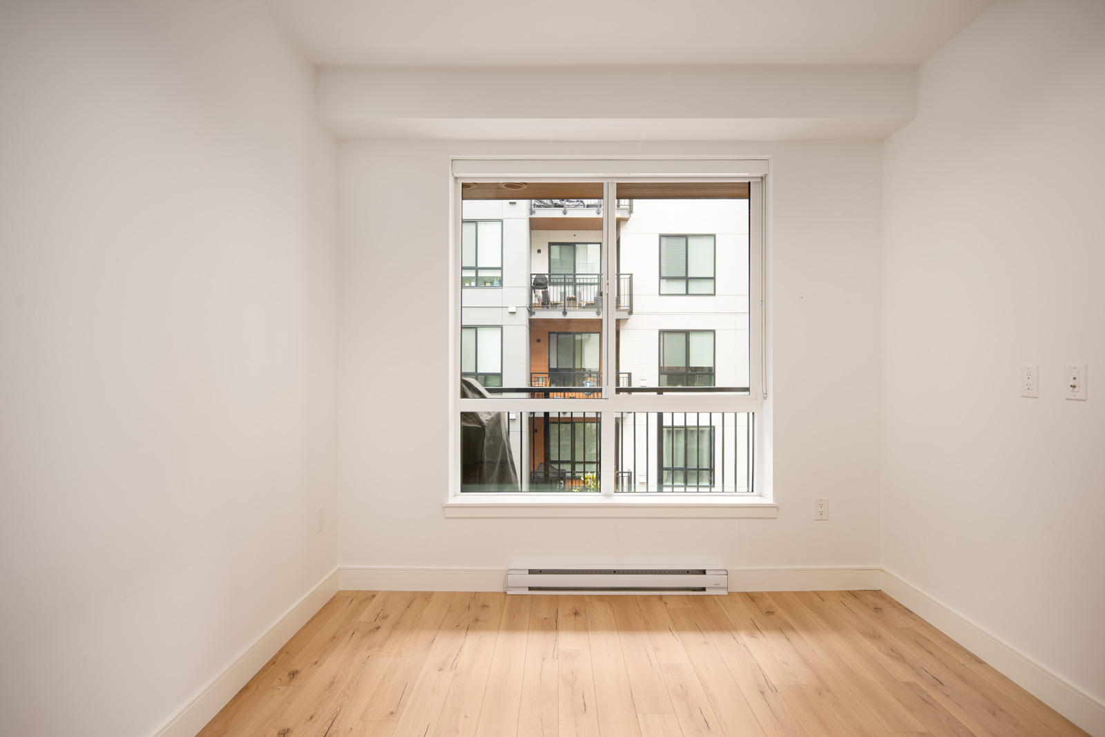 Empty room with light wood flooring, white walls, and a large window overlooking a neighboring apartment building.