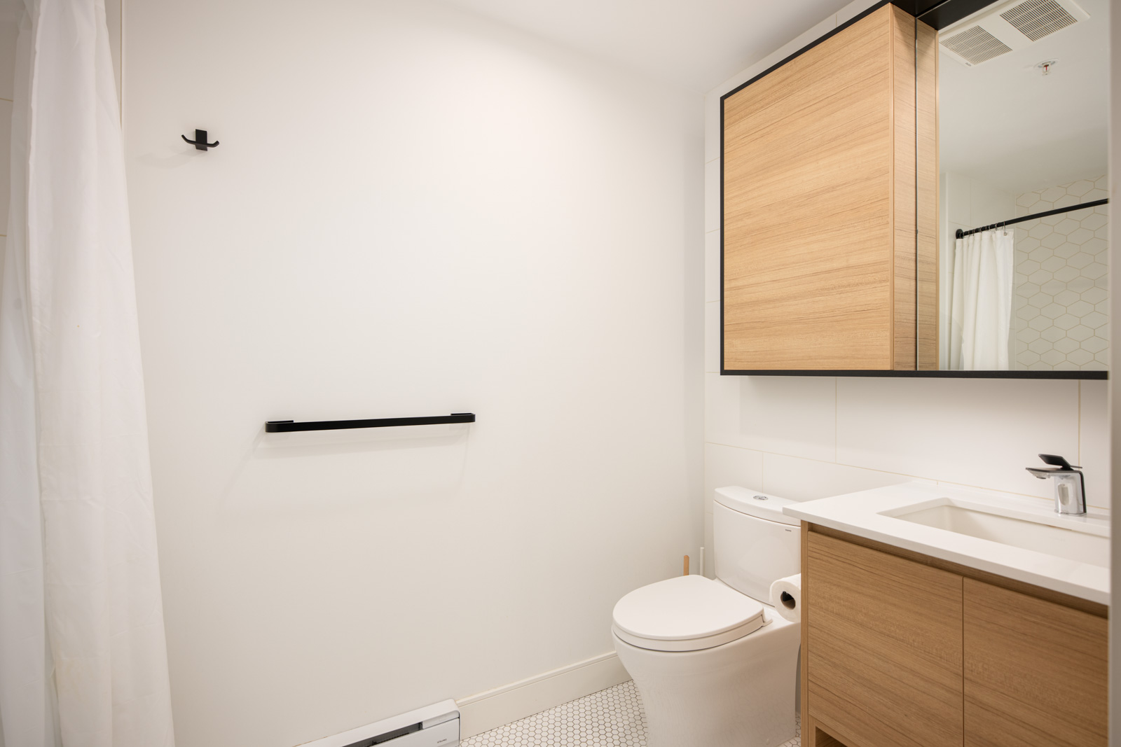 Modern bathroom with light wood cabinets, white walls, a countertop sink, toilet, black towel rail, and a shower with a white curtain.