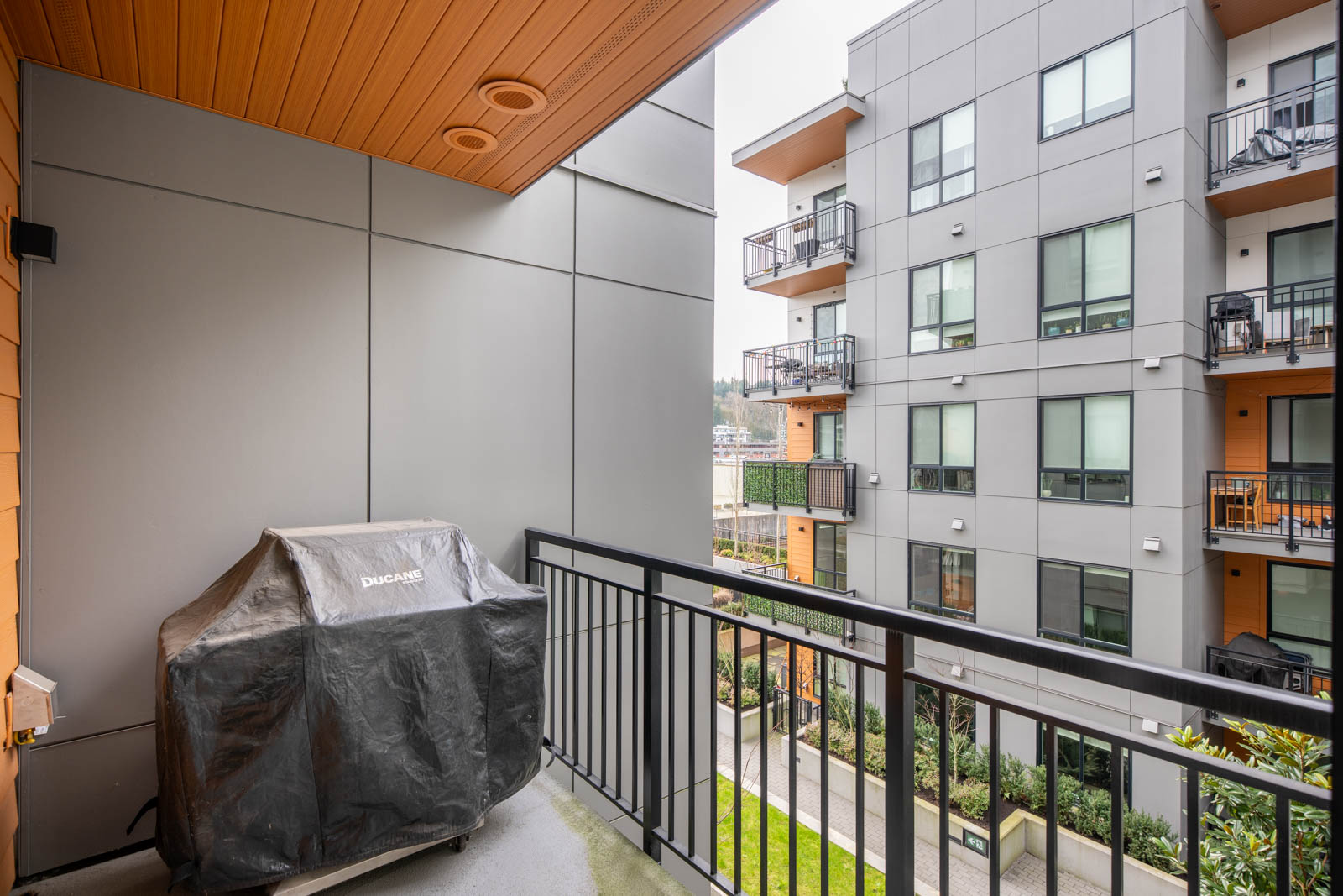 A covered grill sits on a small apartment balcony with metal railings, overlooking a modern multi-story building and landscaped courtyard.