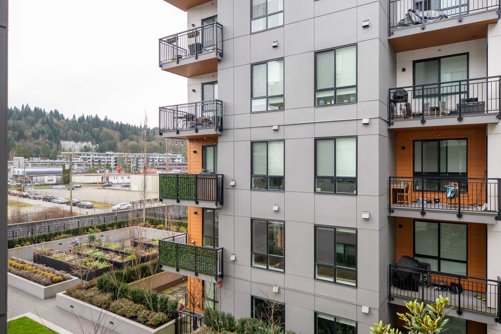 A modern apartment building with balconies, large windows, and a courtyard garden; a commercial area and forested hills are visible in the background.