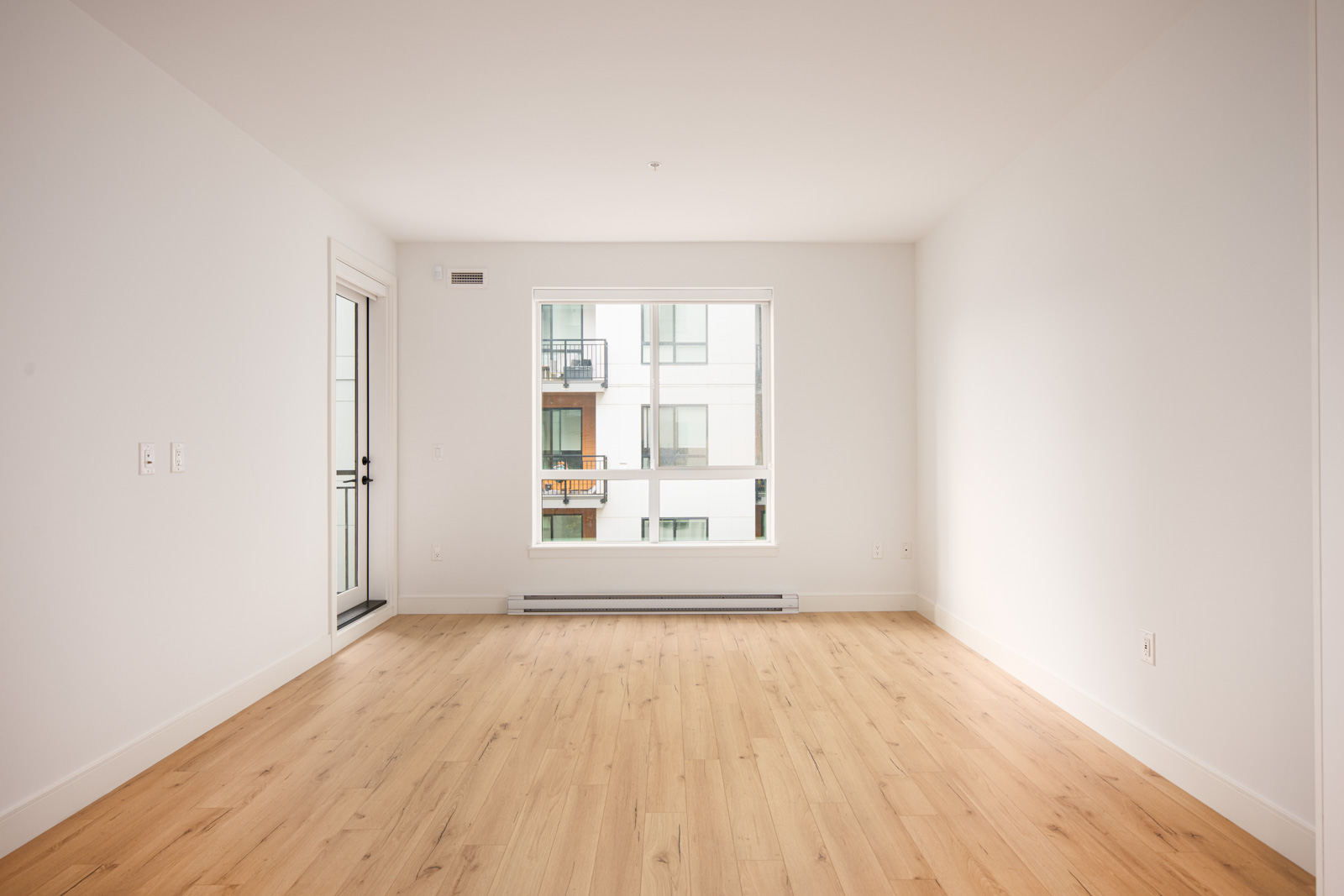 Empty room with light wood flooring, white walls, a large window, and a door leading to a balcony. An apartment building is visible through the window.