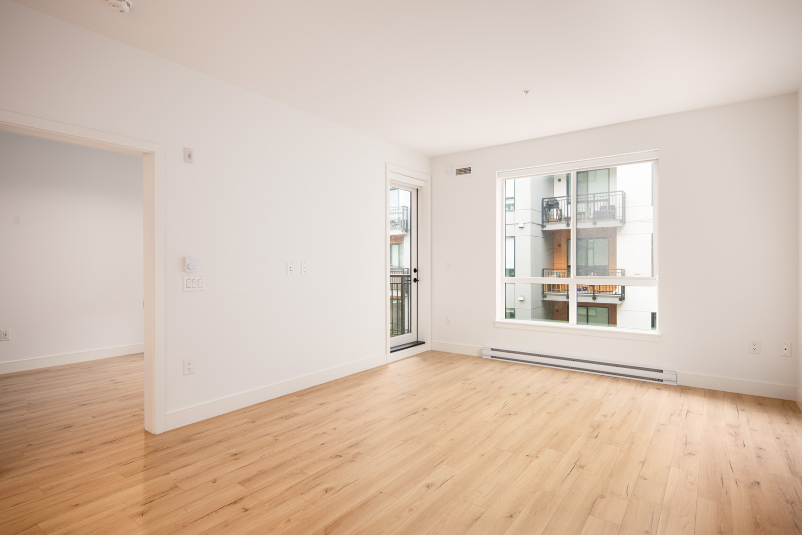 Empty modern room with light wood flooring, white walls, large window, glass door to balcony, and view of an apartment building exterior.