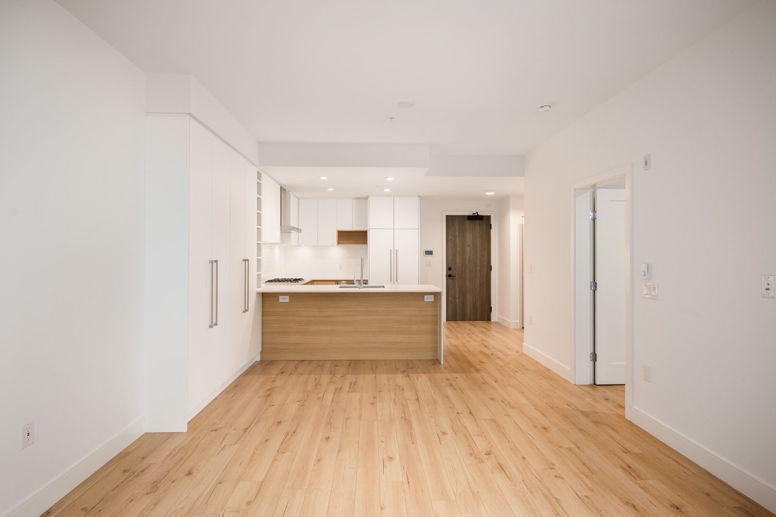A modern, unfurnished apartment interior with light wood flooring, white walls, an open kitchen with a wood island, and a hallway leading to a door.