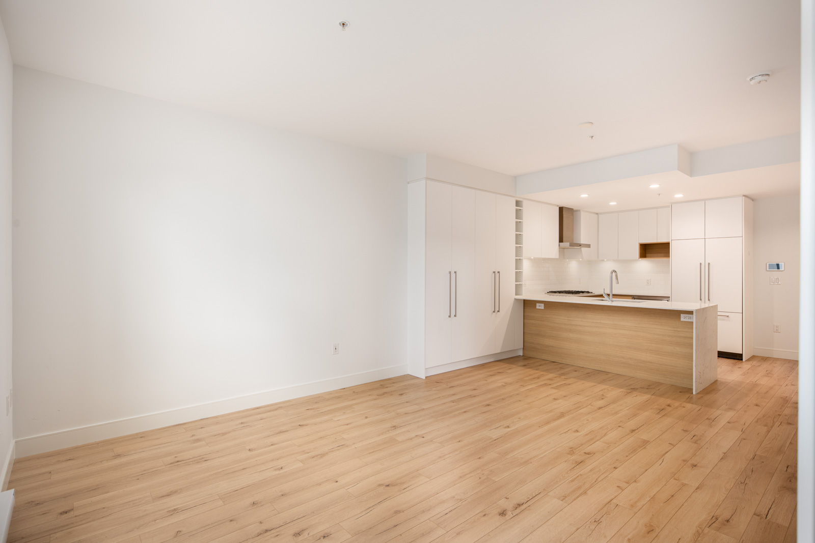 Modern empty apartment with light wood flooring, white walls, and a kitchen featuring white cabinets, built-in appliances, and a wood-accented island with a sink.