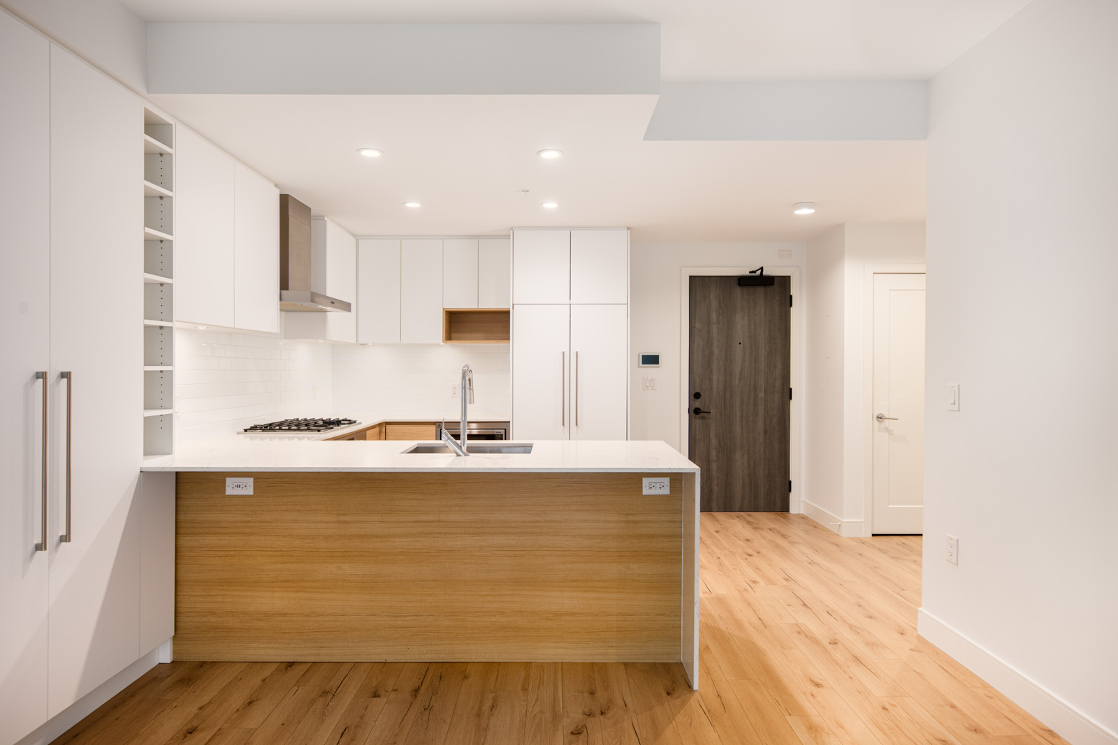 Modern kitchen with white cabinets, wood accents, an island countertop with sink, built-in stove, white backsplash, and light wood flooring. Doorways are visible in the background.